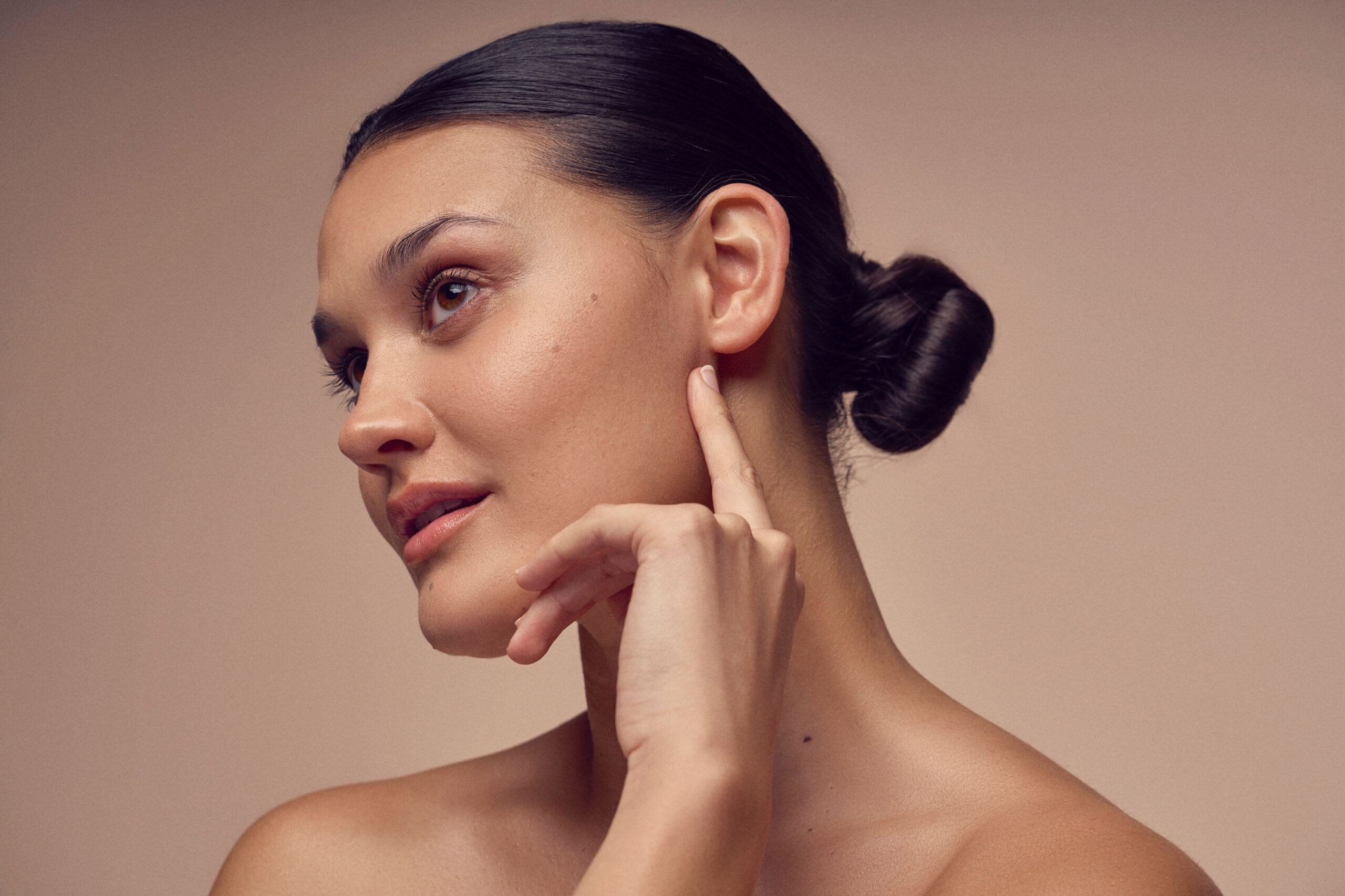 A person with dark hair in a bun looks to the side, gently touching their face with one hand. The background is a neutral tone.