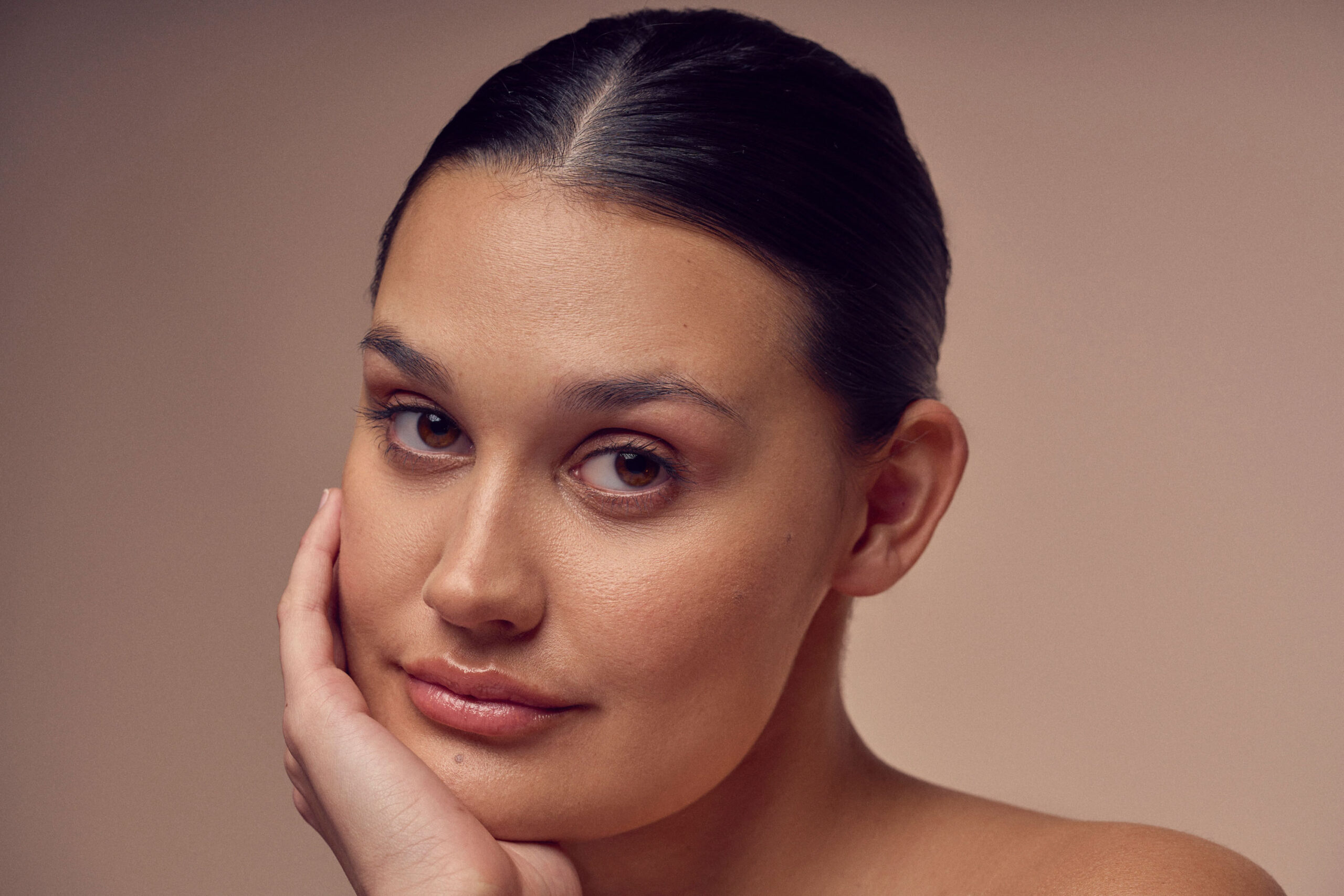 A person with dark hair pulled back, gazing forward with a neutral expression. They rest their chin on one hand, with a plain background that emphasizes their serene look.