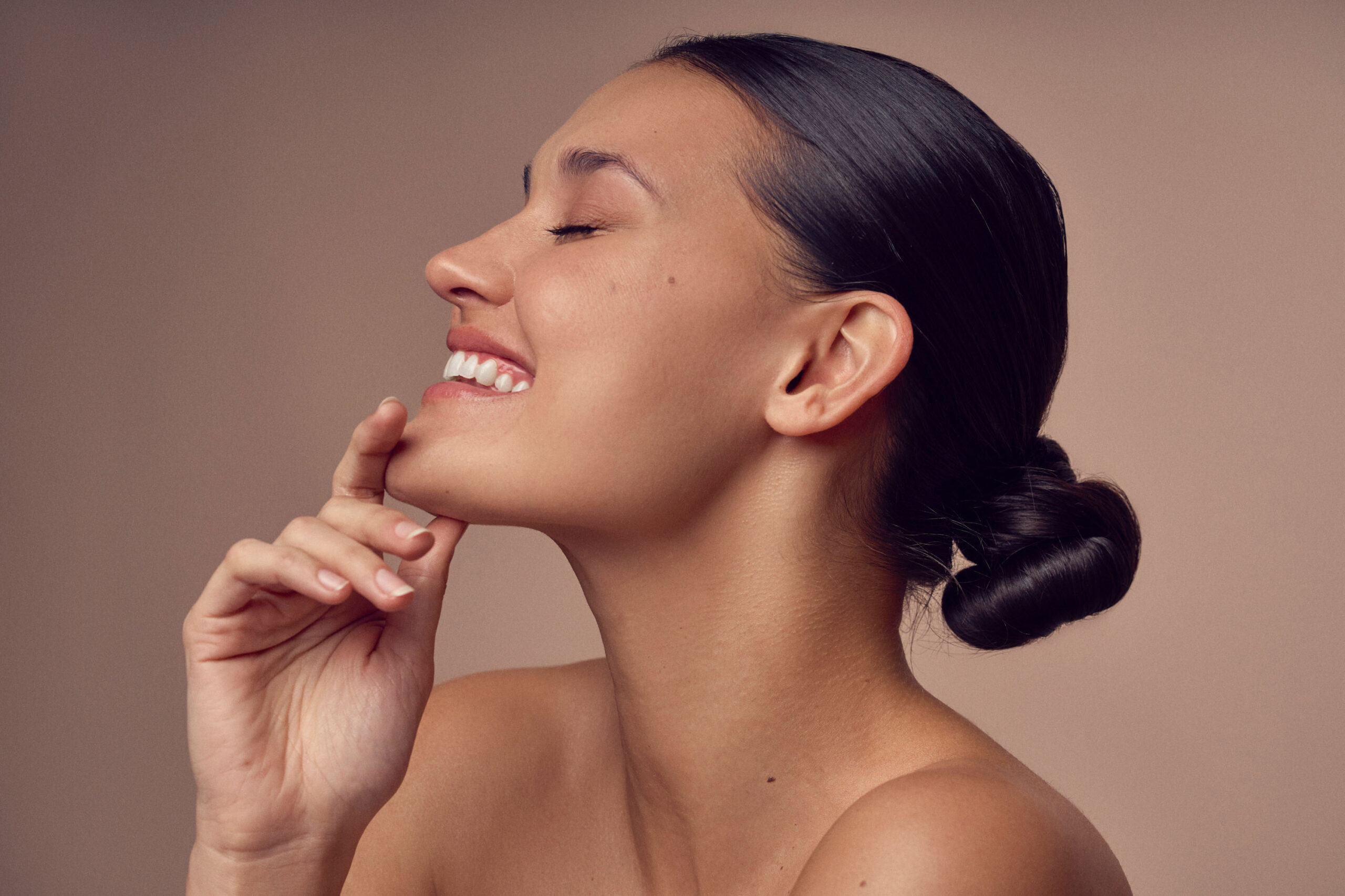 A woman with smooth skin smiles gently in profile against a beige background. Her hair is neatly tied back in a bun, and she is touching her chin with her fingers, exuding a sense of calm and contentment.