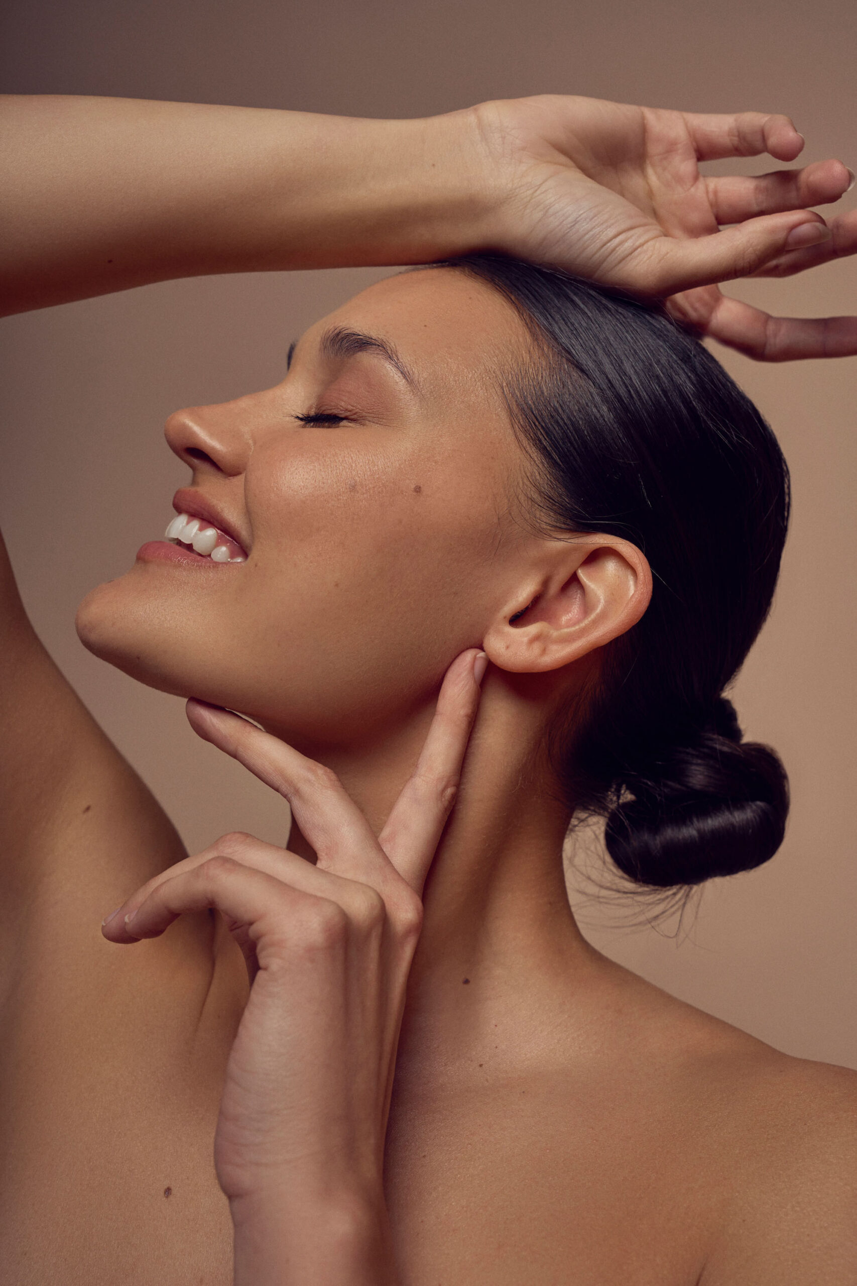 A woman with smooth skin poses with her eyes closed, one arm over her head and the other hand touching her neck. Her dark hair is styled in a bun. The background is neutral, enhancing the serene and elegant mood.