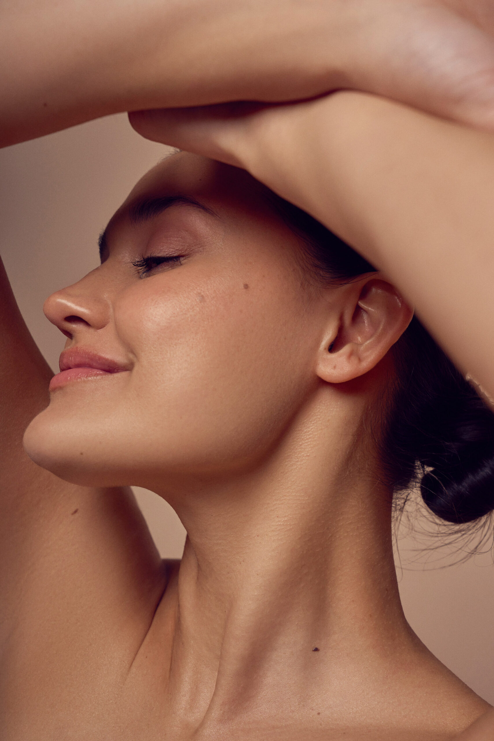 A woman with her eyes closed and a serene expression tilts her head back, with one arm raised above her head. Her skin is glowing and smooth, and her dark hair is pulled back, leaving her face unobstructed.