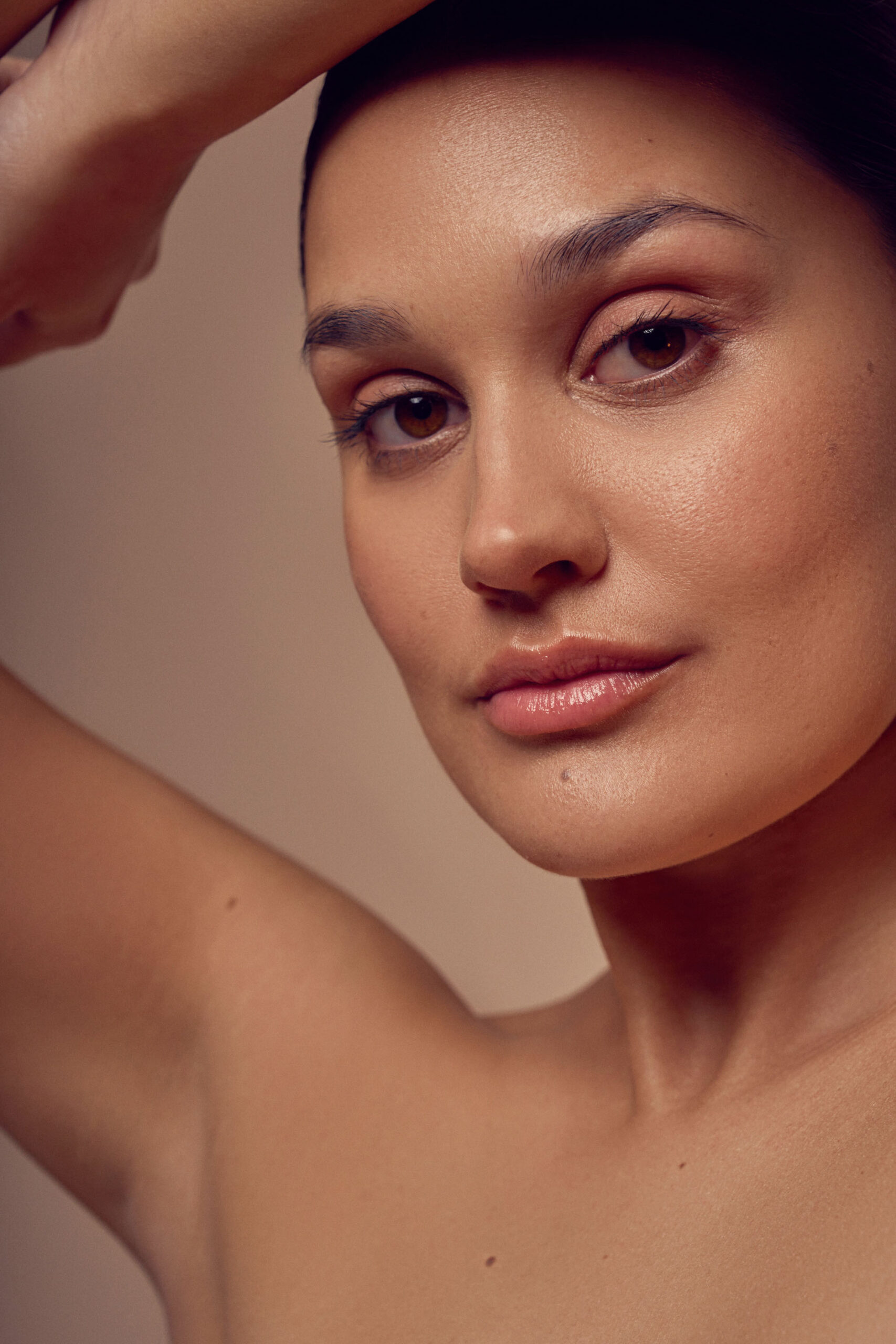 A close-up portrait of a person with a calm expression. They have smooth skin, subtly arched eyebrows, and slightly parted lips. Their left arm is raised above their head against a neutral background.