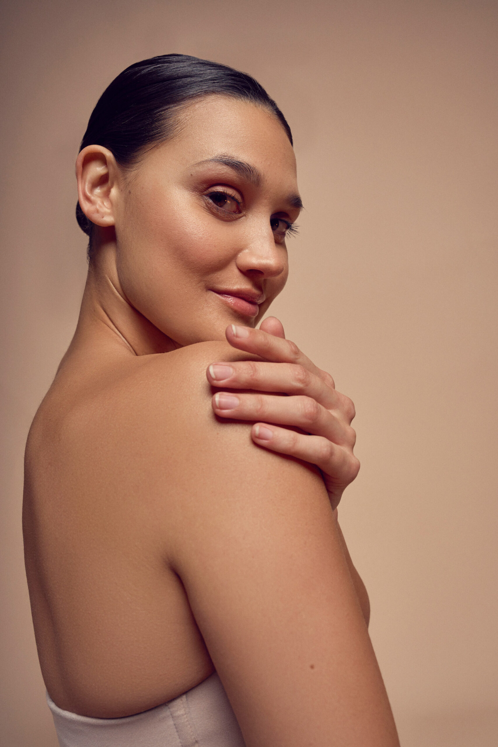 A woman with a sleek hairstyle poses against a beige background. She is looking over her bare shoulder with a soft smile, gently touching her shoulder with one hand. Her posture is elegant and confident.
