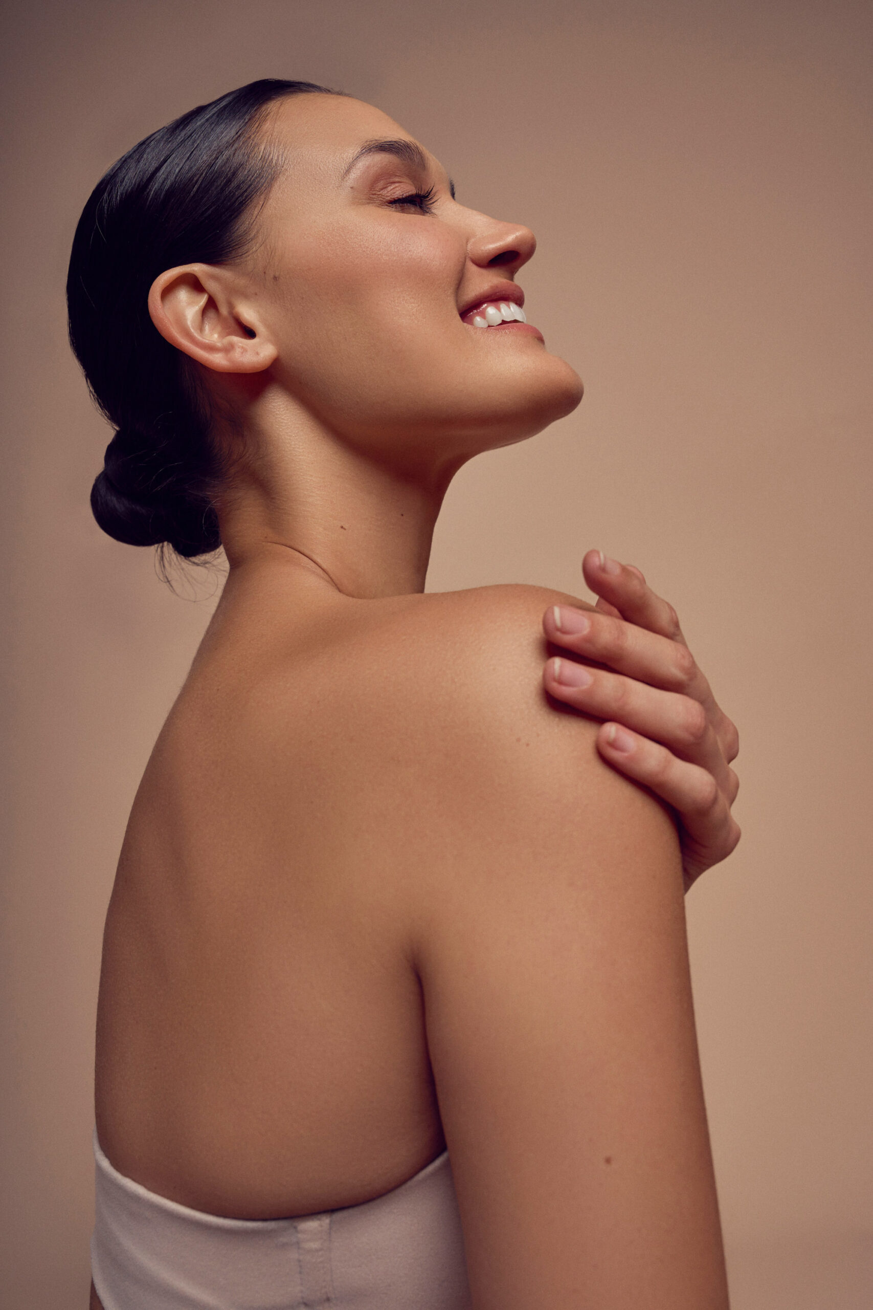 A woman with dark hair in a bun smiles with her eyes closed. Her head is turned to the side, and she lightly places one hand on her shoulder. She wears a light-colored strapless top against a neutral background.