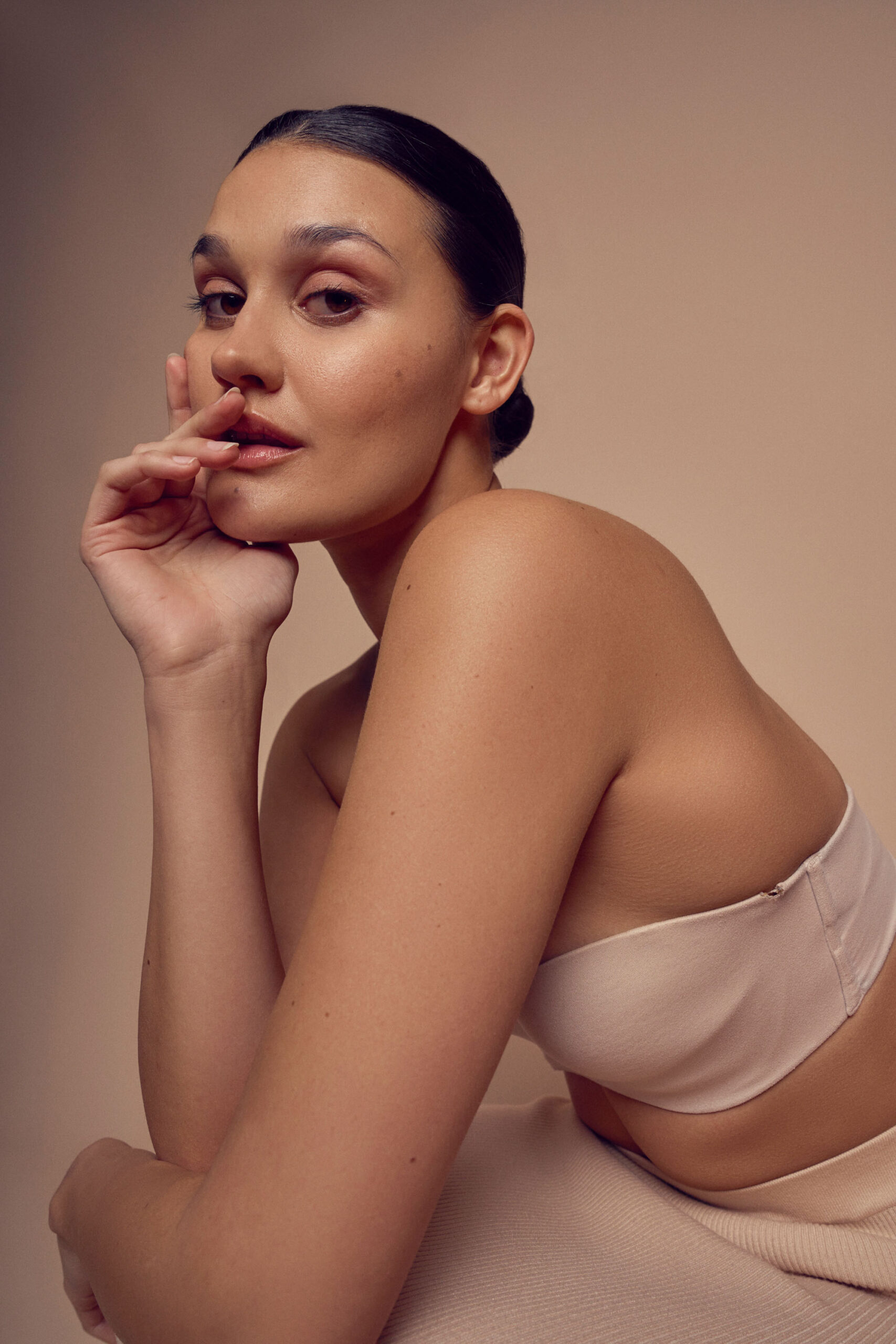 A woman with dark hair, styled in a bun, sits with her elbow resting on her knee and her fingers gently touching her lips. Shes wearing a beige strapless top and looks thoughtfully to the side against a neutral background.