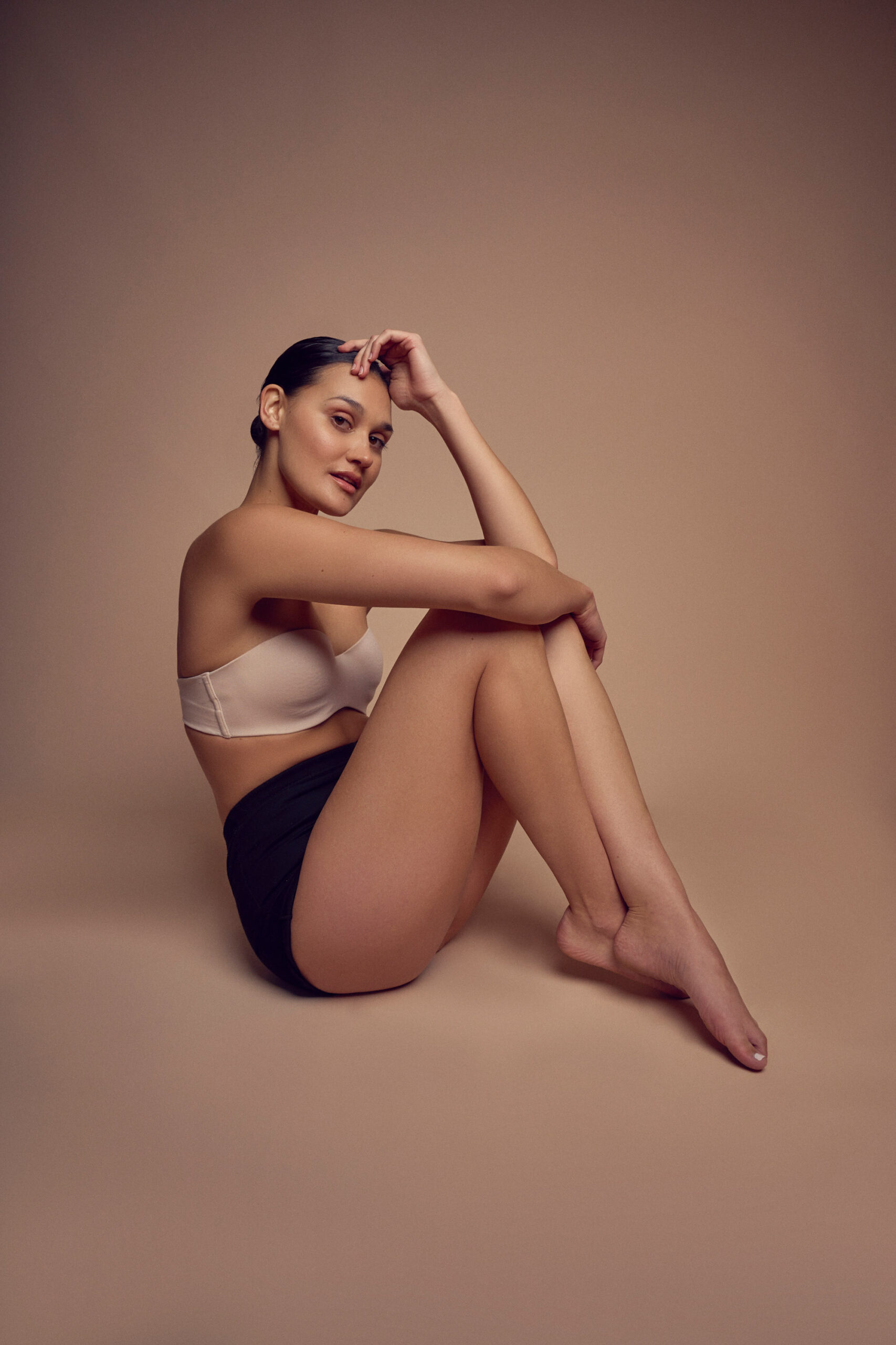 A woman sits on a smooth, neutral-toned backdrop with her knees drawn up, wearing a light-colored bra and dark shorts. Her head is tilted slightly, right arm resting on her knee, and she gazes softly forward.