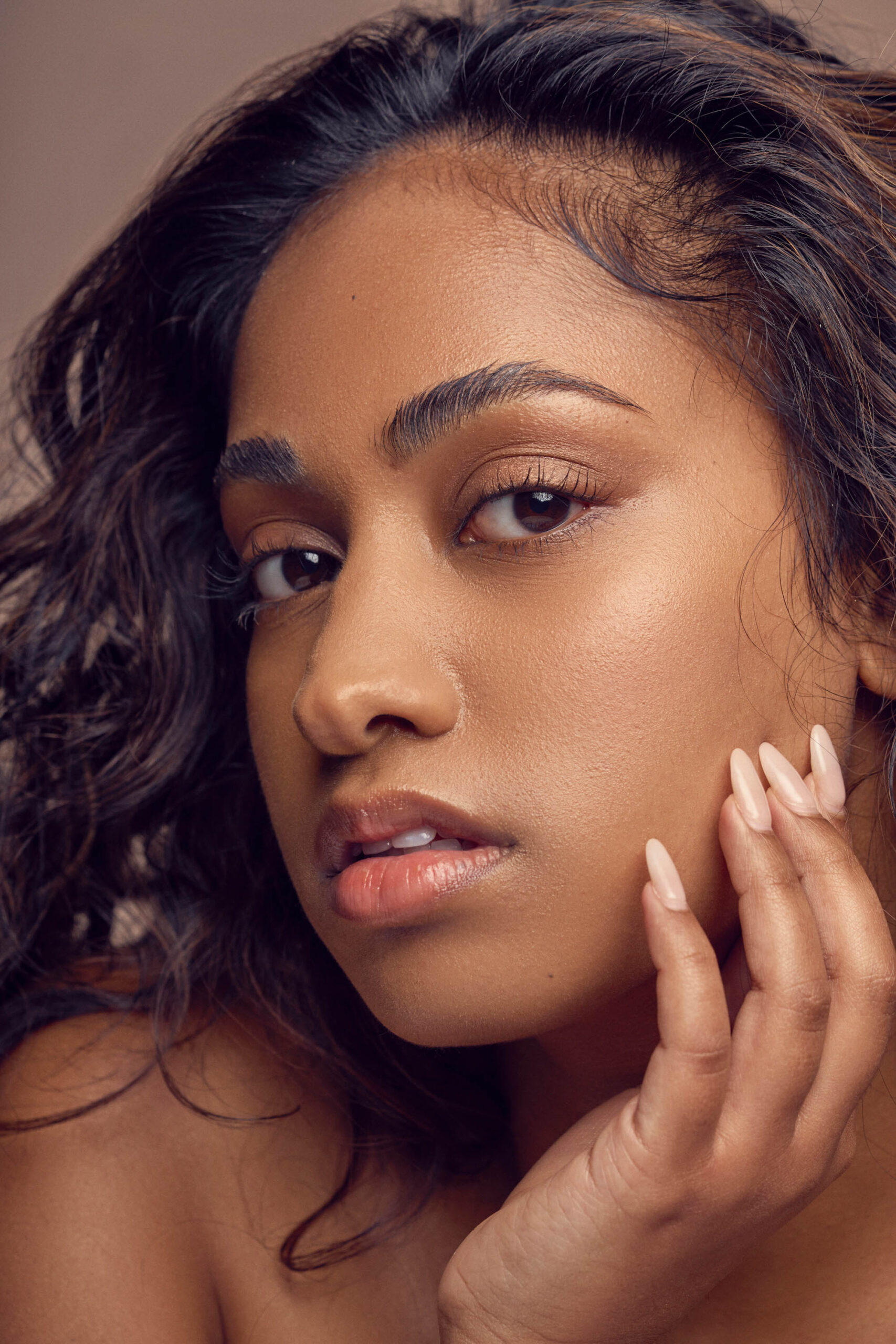 A person with long, wavy hair and smooth skin poses with their hand touching their cheek, looking directly at the camera against a neutral background. Their expression is serene and contemplative.