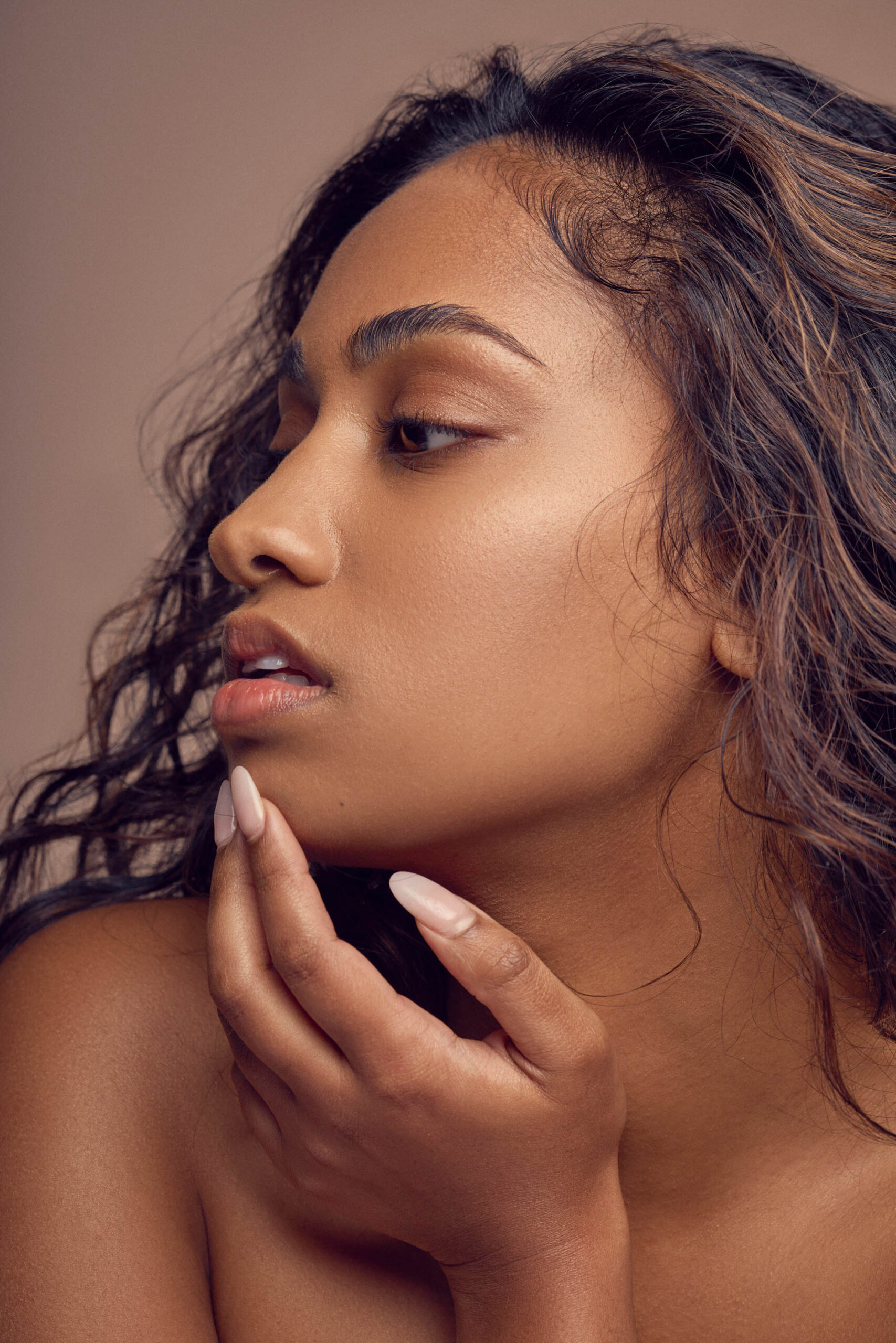 A woman with long, wavy hair gazes to the side. Her hand softly touches her chin. The background is a neutral shade, and the lighting highlights her smooth skin and facial features.