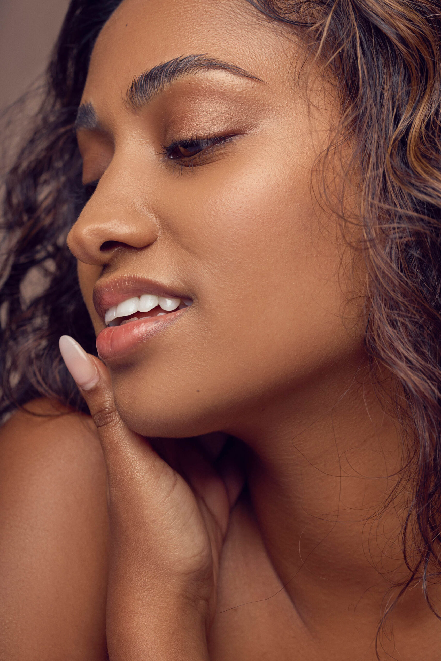 Close-up of a person with wavy hair, softly smiling with eyes closed. They gently touch their cheek with manicured fingers. Their skin appears smooth and radiant, and the background is neutral, highlighting their serene expression.