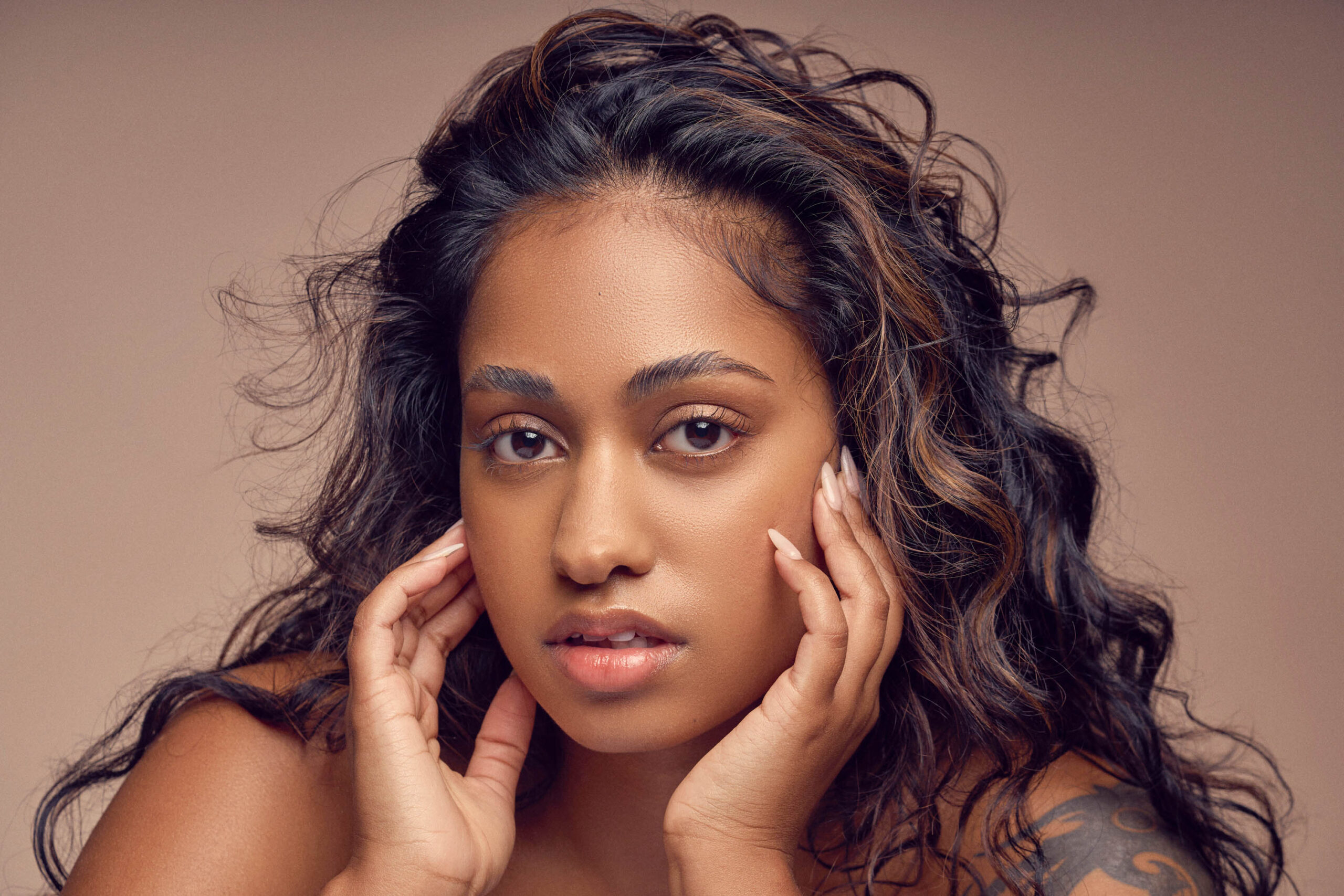 A person with long, wavy hair gently touches their face with both hands. They have a contemplative expression and are posed against a neutral background.