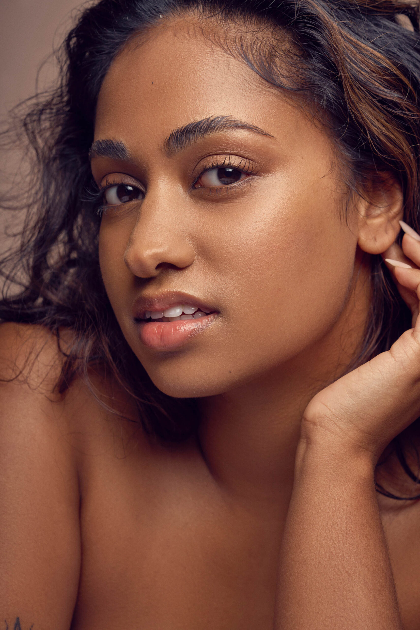 Close-up portrait of a person with long, wavy hair and glowing skin. They are looking at the camera with a neutral expression, their hand gently touching their face. The lighting highlights their features and creates a warm, soft tone.