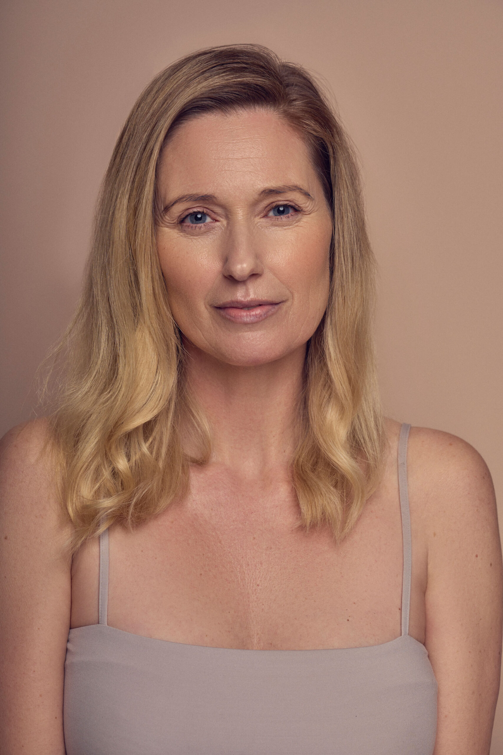 A woman with long blond hair and a neutral expression is wearing a sleeveless top against a beige background.