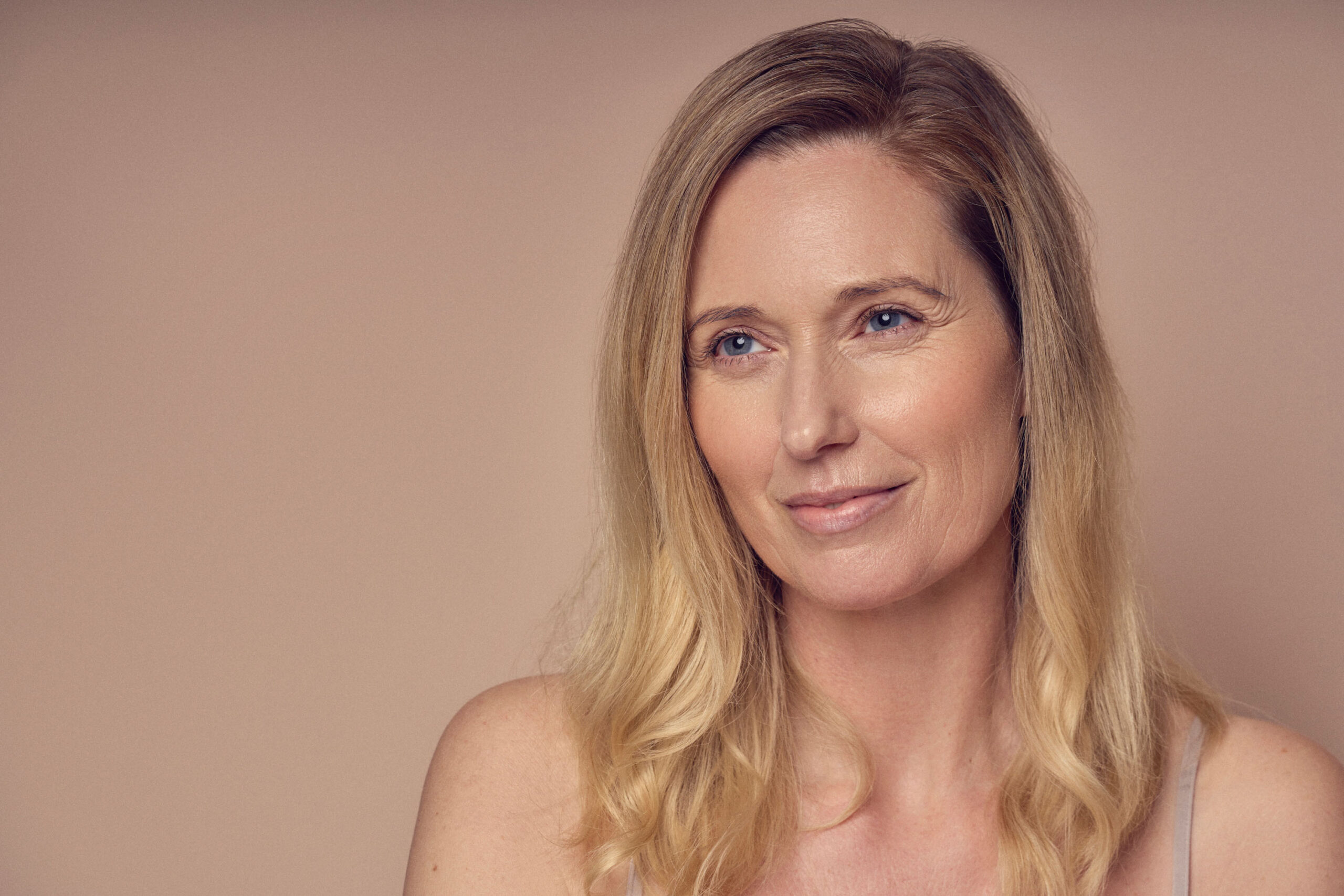 A woman with long blonde hair and wearing a sleeveless top looks slightly to the side with a gentle smile against a neutral background.