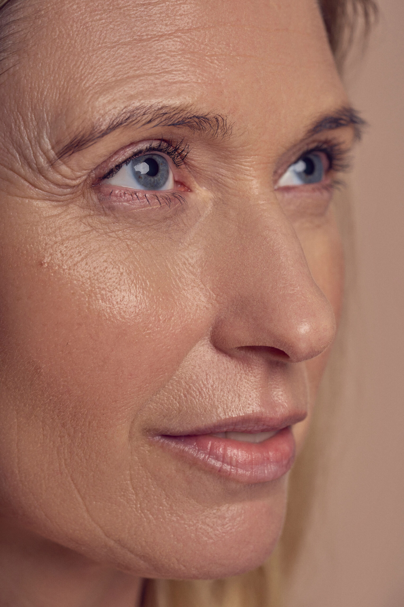 Close-up of a persons face focusing on their eyes and part of their nose and mouth. The skin texture is visible, highlighting natural details and light reflections. The background is softly blurred in a neutral tone.