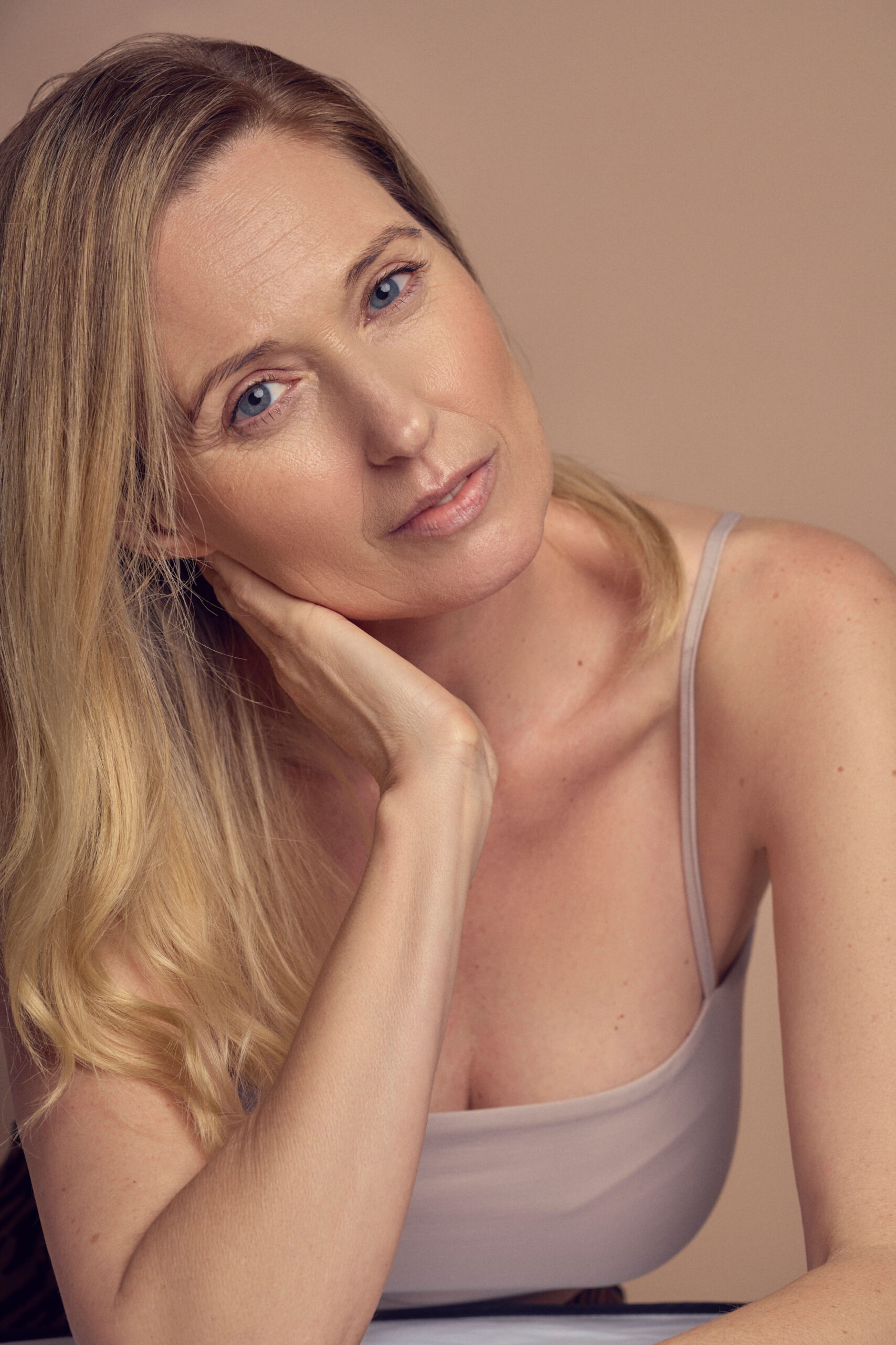 A woman with long blonde hair and blue eyes is resting her chin on her hand. She is wearing a sleeveless top and is looking softly at the camera against a neutral background.
