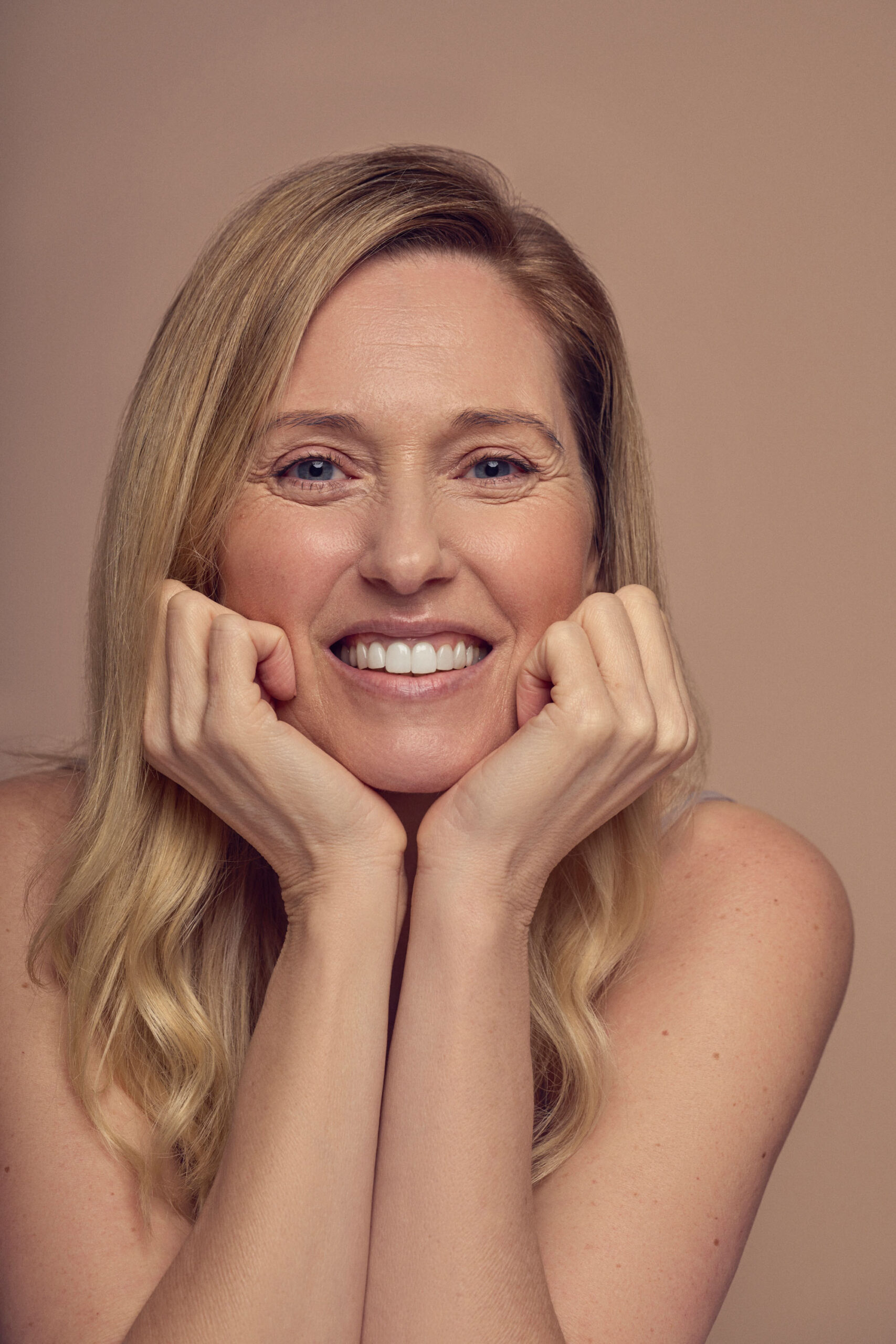 A smiling woman with long, blonde hair rests her chin on her hands. She is gazing directly at the camera against a plain beige background.