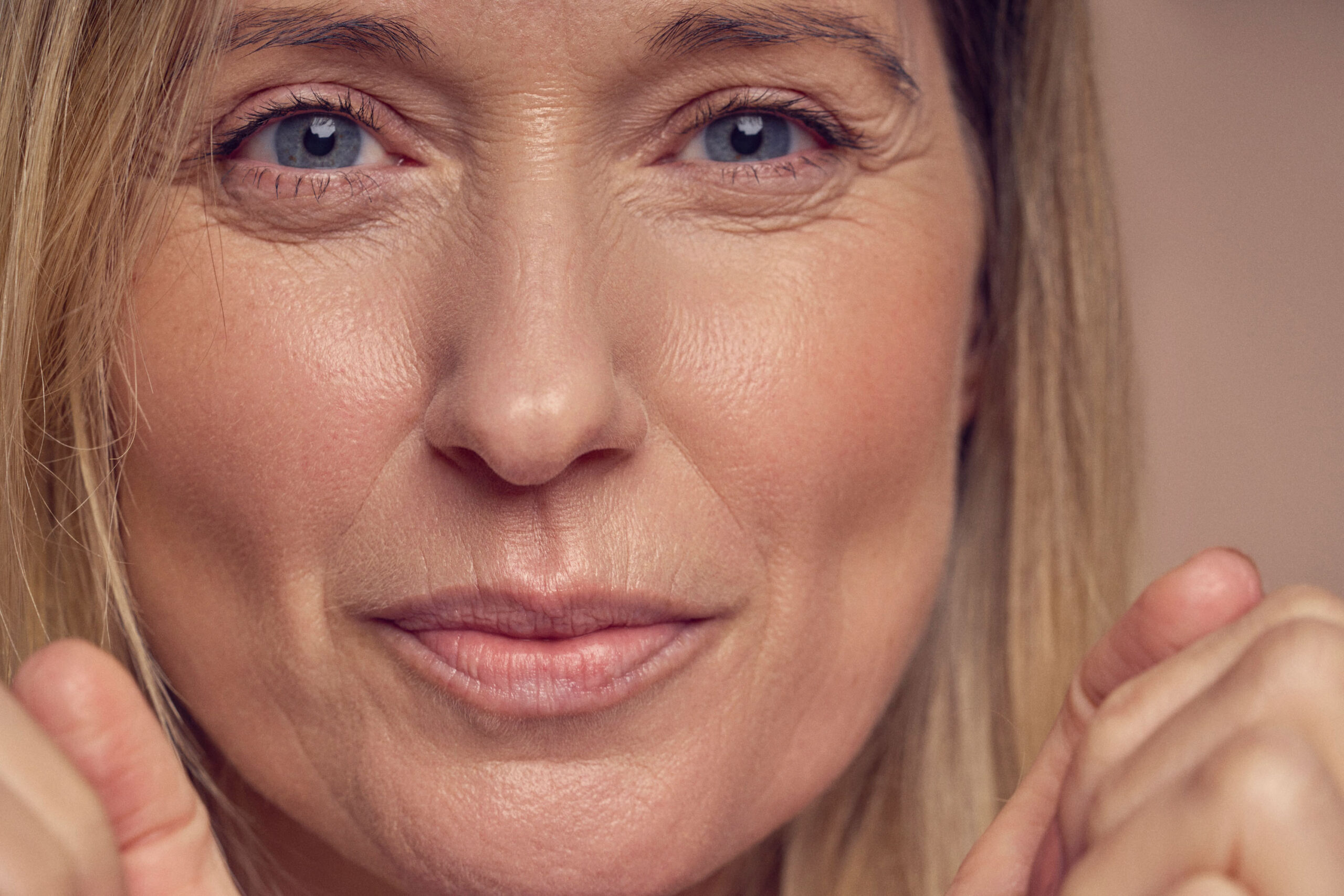 Close-up of a woman with light hair, smiling gently. Her hands are partially visible in the foreground, suggesting a relaxed pose. The image focuses on her face, highlighting her eyes and natural expression.