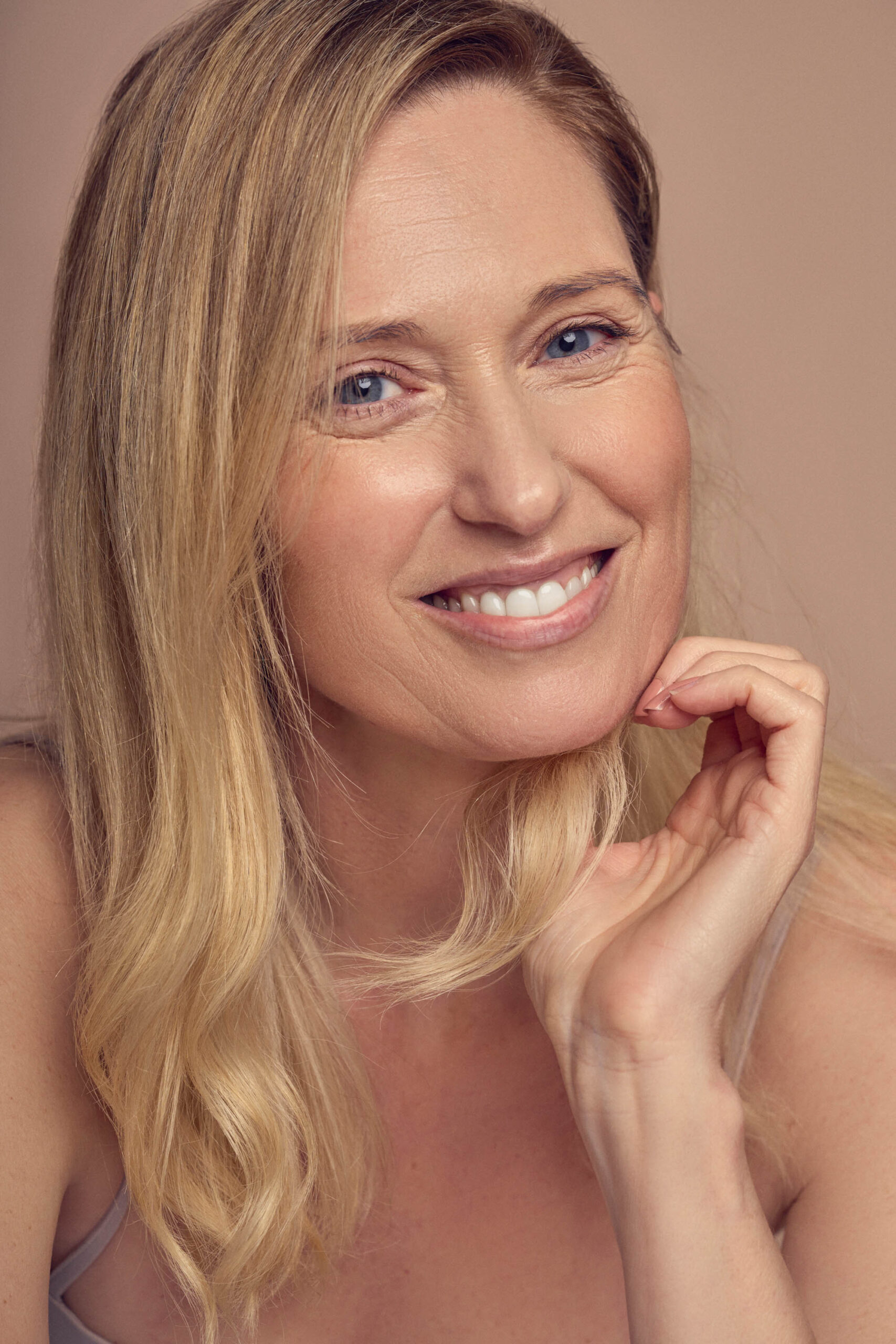 A smiling woman with long blonde hair poses against a neutral background. She rests her chin lightly on her hand, displaying a natural and relaxed expression.