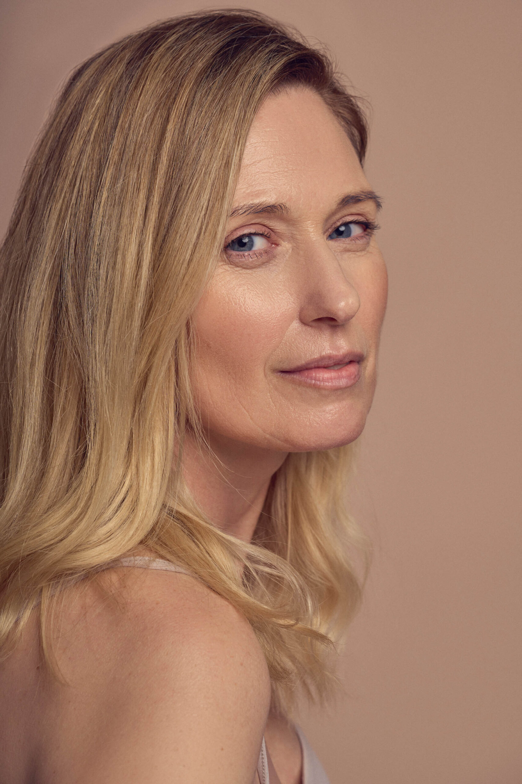 A woman with long blonde hair and blue eyes looks over her shoulder, set against a neutral background. She has a soft expression and a subtle smile, wearing a sleeveless top.