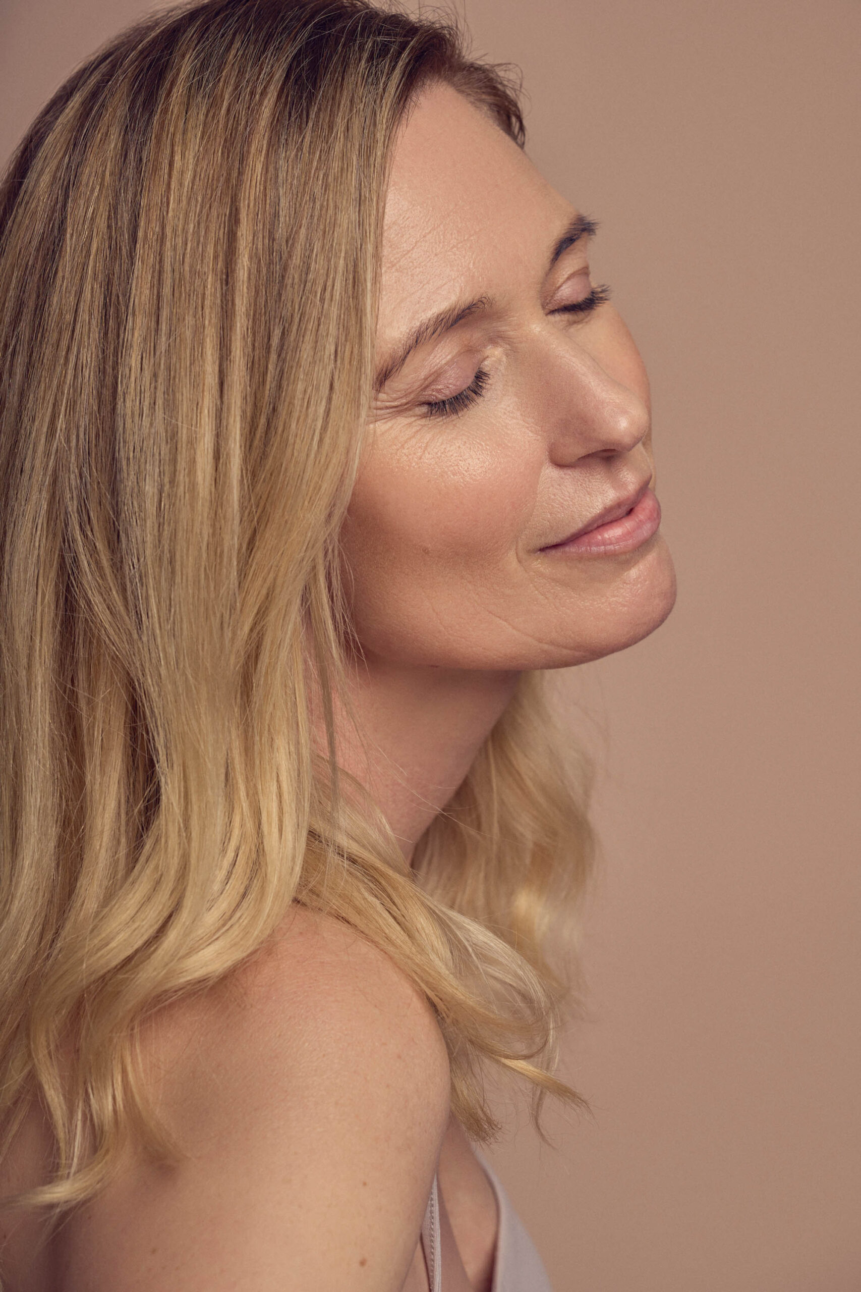 A woman with long blond hair peacefully smiles with her eyes closed against a soft beige background. Her head is slightly tilted back, and she is wearing a sleeveless top.