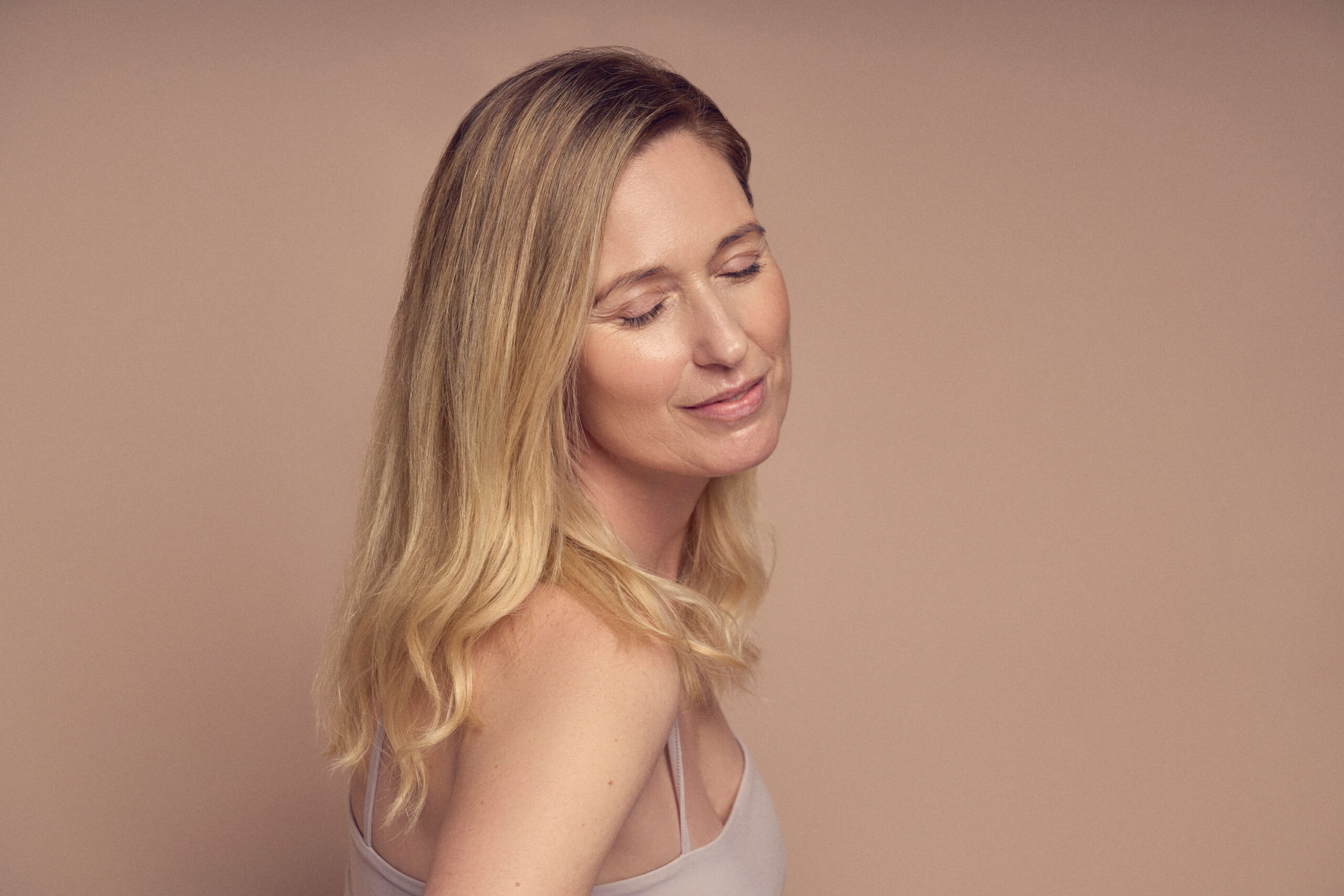 A woman with long blonde hair and light skin smiles gently with her eyes closed against a soft beige background. She is wearing a simple, light-colored top and appears relaxed and content.