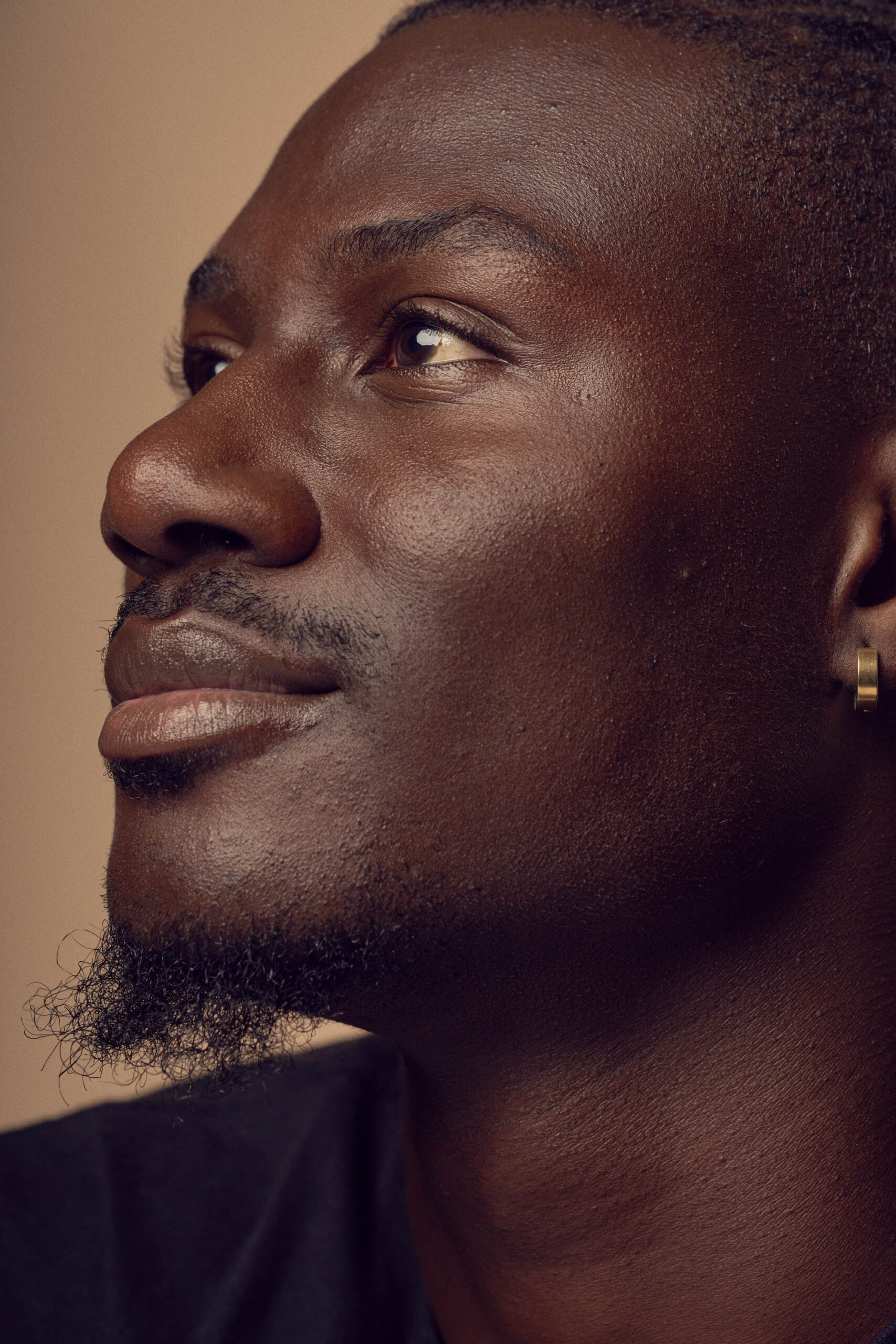 A close-up profile of a person with short hair and a goatee, looking up thoughtfully. They have an earring and a slight smile, set against a neutral background.