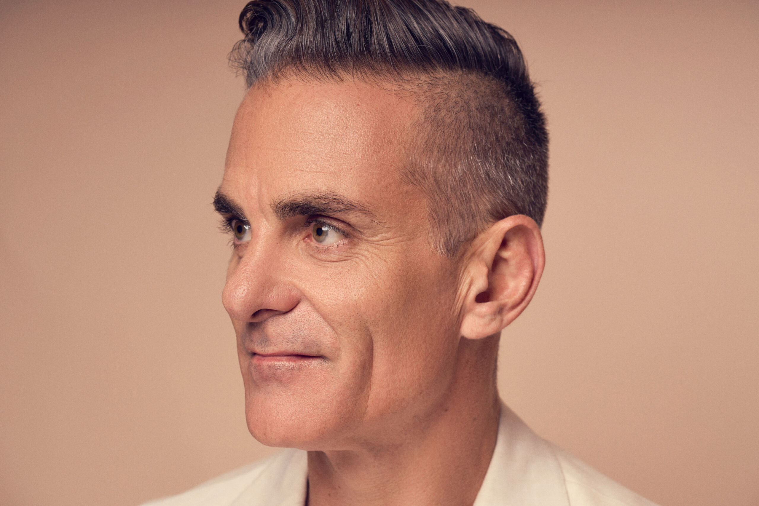 A man with short, styled gray hair and a clean-shaven face is looking to the side against a beige background. He wears a light-colored shirt and has a calm expression.