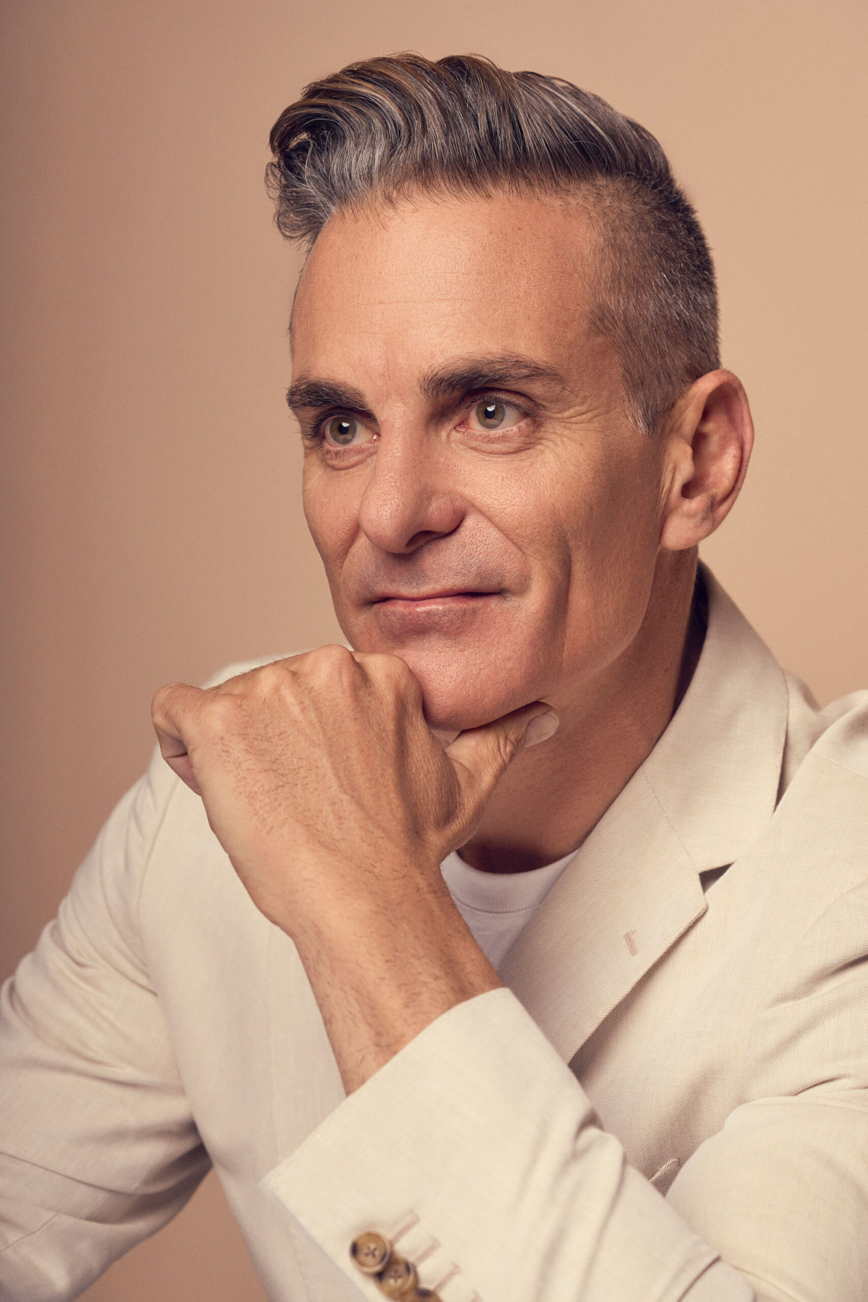 A man with short, gray hair and a slight smile is wearing a beige blazer over a white shirt. He rests his chin on his hand while gazing thoughtfully. The background is a soft beige color.