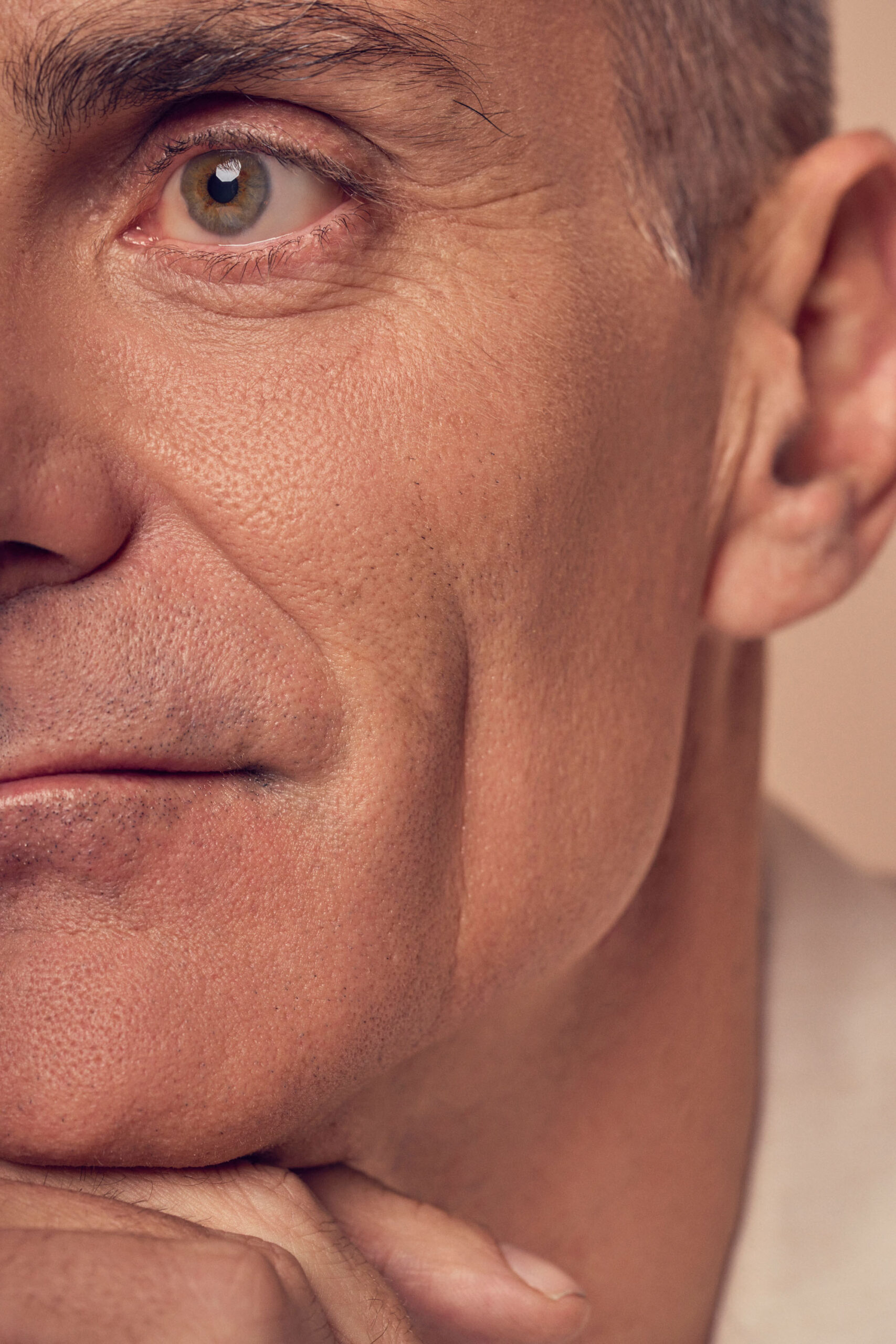 Close-up of a middle-aged man’s face, focusing on the left eye and cheek. He has short hair and rests his chin on his hand. The image captures detailed skin texture and expression in a soft, neutral background.