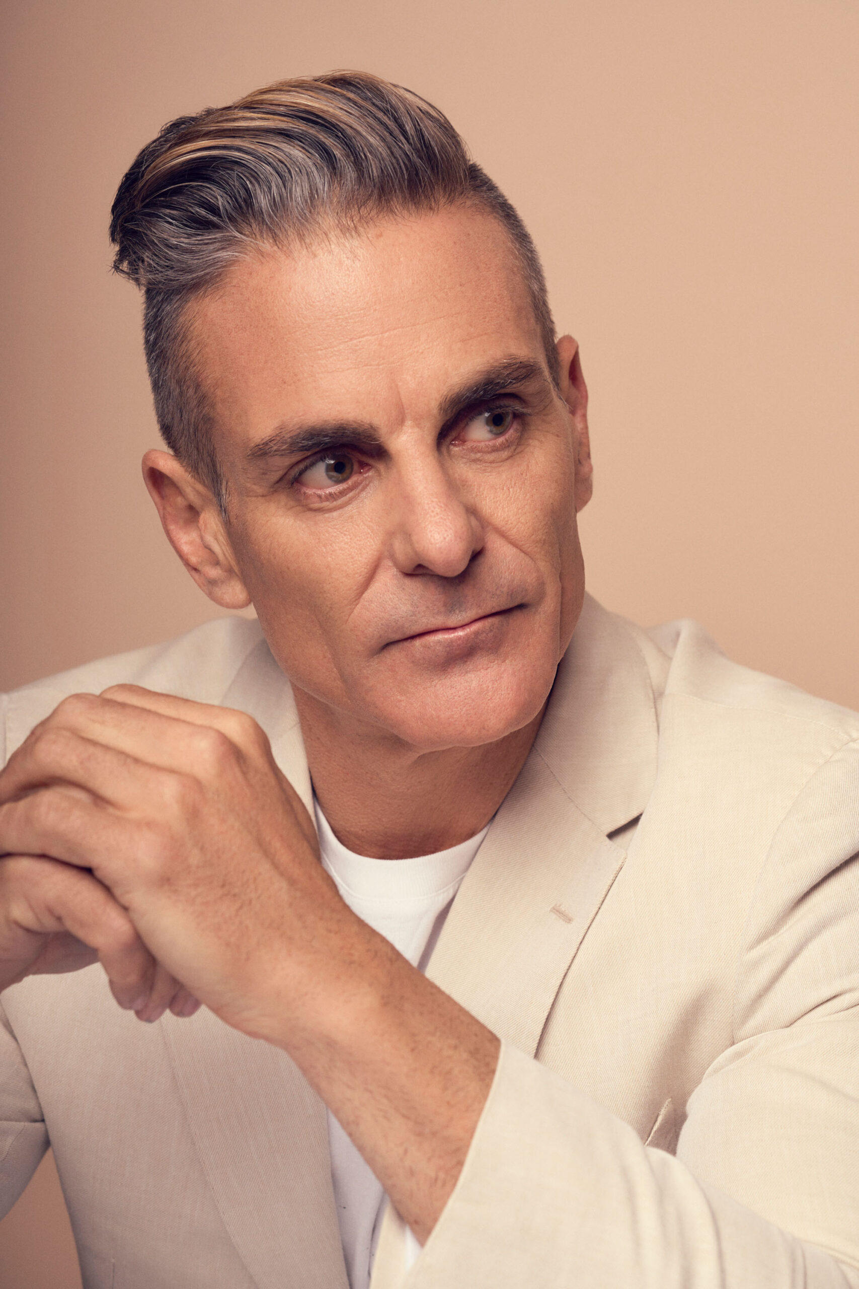 A man with short, slicked-back hair wearing a light beige blazer and white shirt, gazes thoughtfully to the side against a neutral background.