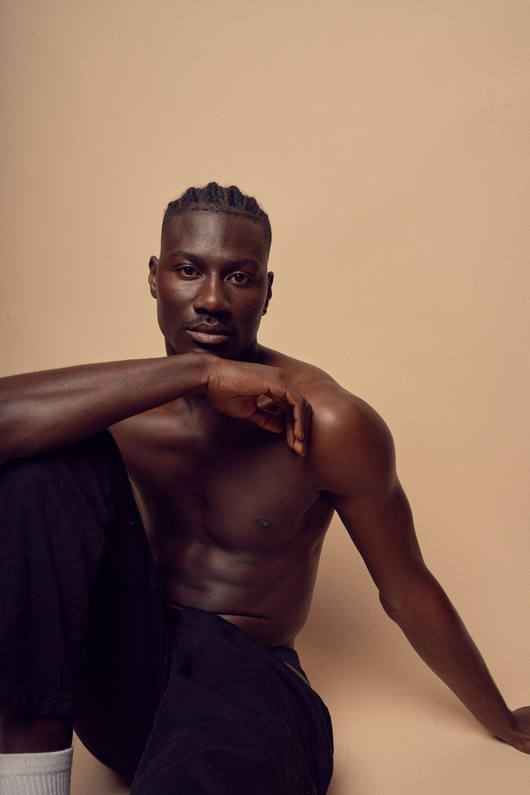 A shirtless man sitting against a beige background, resting one arm on his bent knee while the other arm is extended to the side. He has short braided hair and wears black pants with white socks. His expression is calm and relaxed.