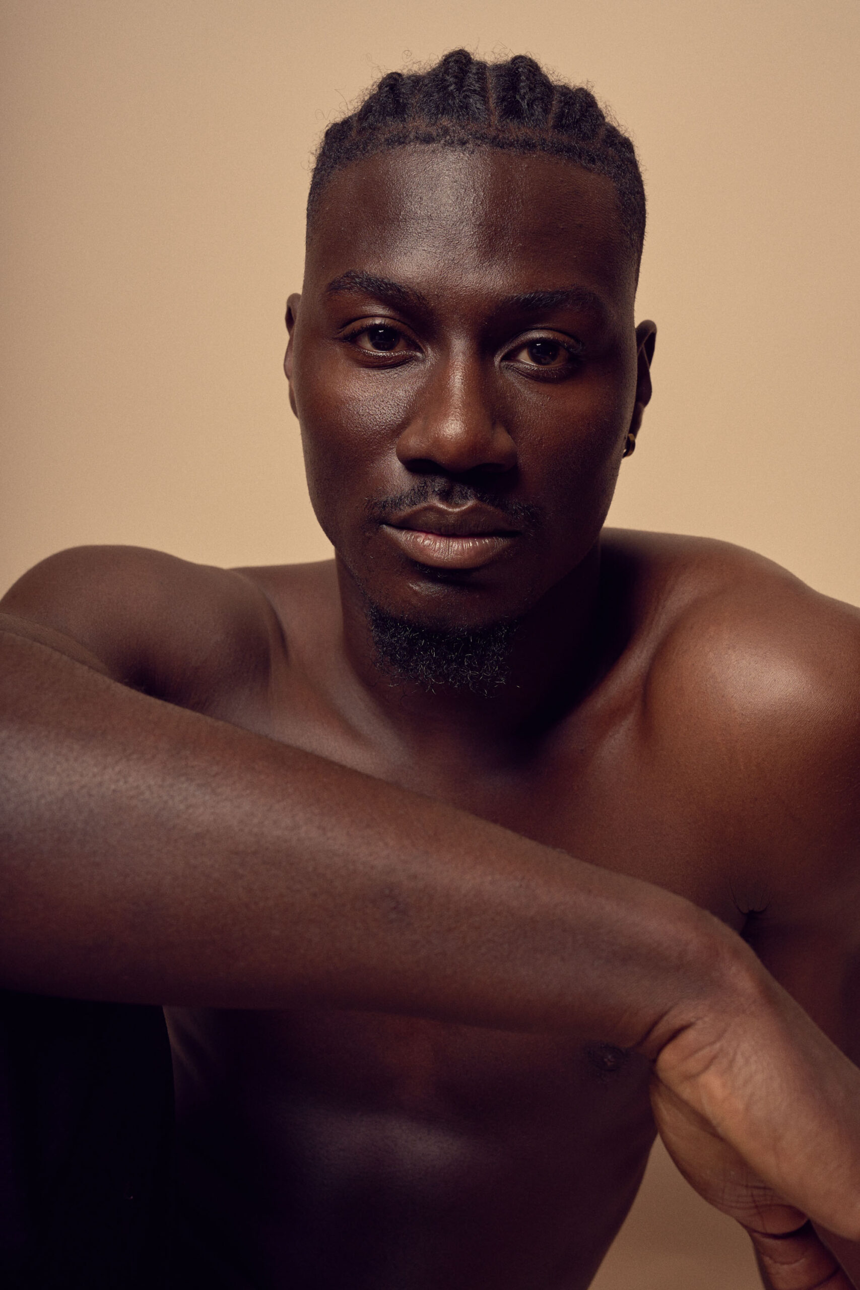 A shirtless man with short braided hair is sitting against a beige background. His arms are resting on his knees, and he is looking directly at the camera with a calm expression.