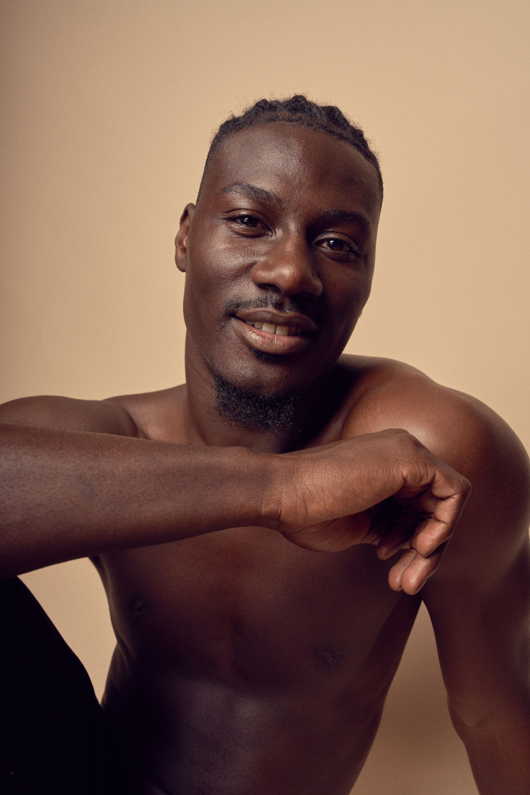 A person with short braided hair and a goatee smiles at the camera. They are shirtless against a neutral beige background, with one arm resting on their knee.