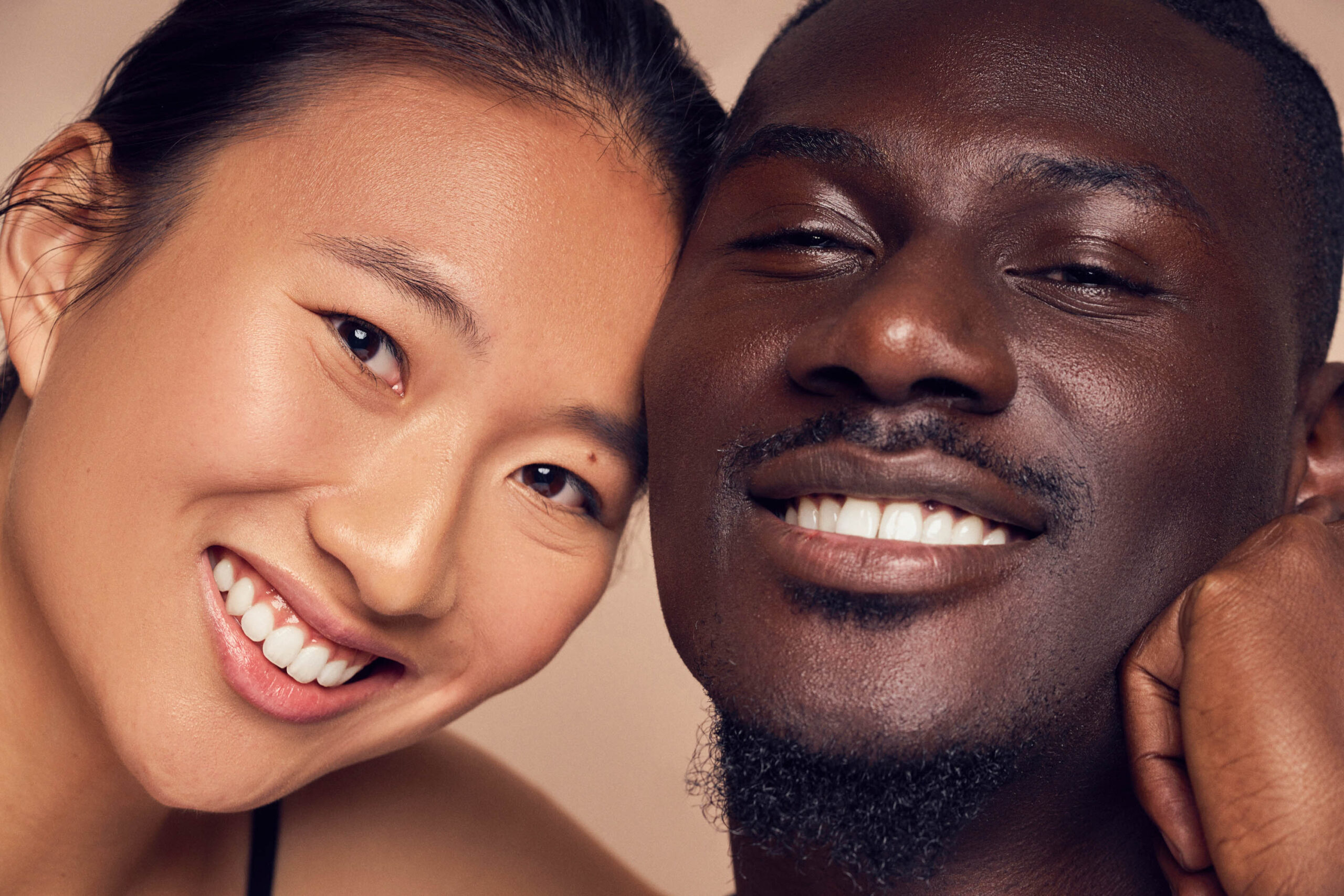 A close-up of two people smiling. One person has long dark hair and the other has short dark hair and a goatee. Both appear to be relaxed and content, with their faces close together against a neutral background.