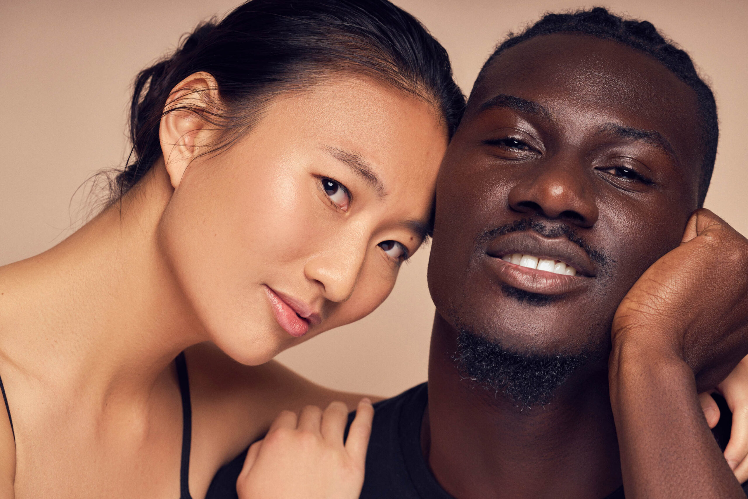 A close-up of a smiling couple, with the woman resting her head on the mans shoulder. The woman has dark hair pulled back, and the man has short black hair. They are both wearing simple, dark tops against a neutral background.