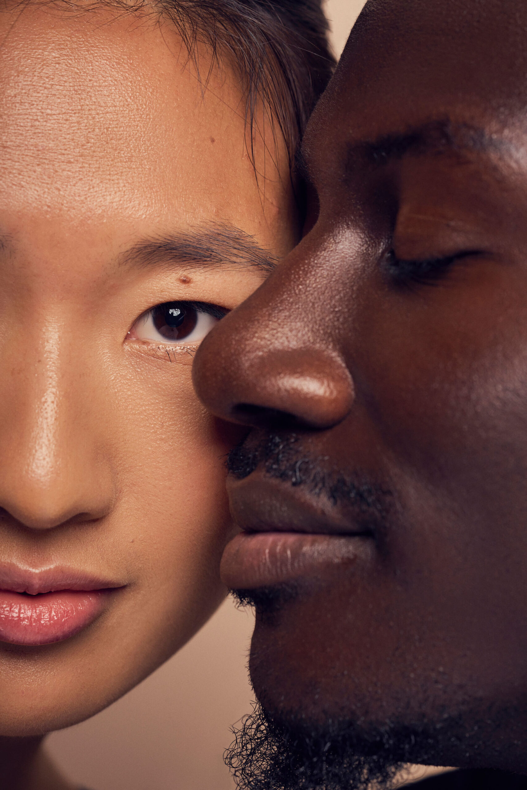 A close-up of two people with their faces touching. One person is facing forward with an eye open, while the other is in profile with eyes closed. The image focuses on the contrasting skin tones and serene expressions.