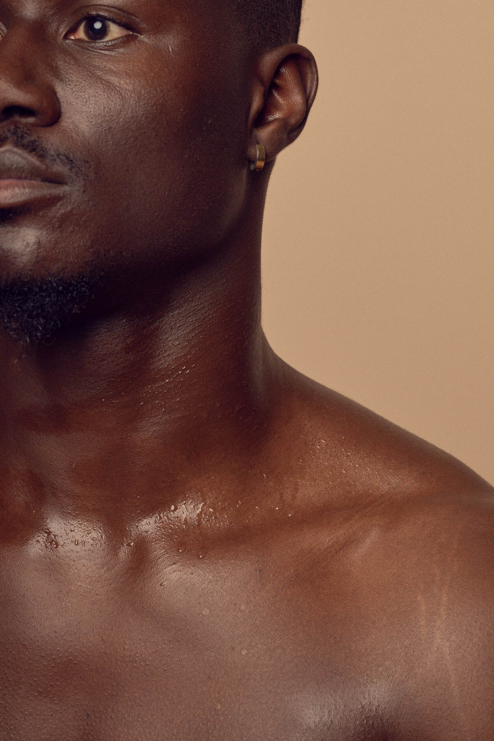 Close-up of a mans face and upper body with smooth dark skin against a beige background. He has a short beard, is wearing a hoop earring, and his skin glistens under the light.