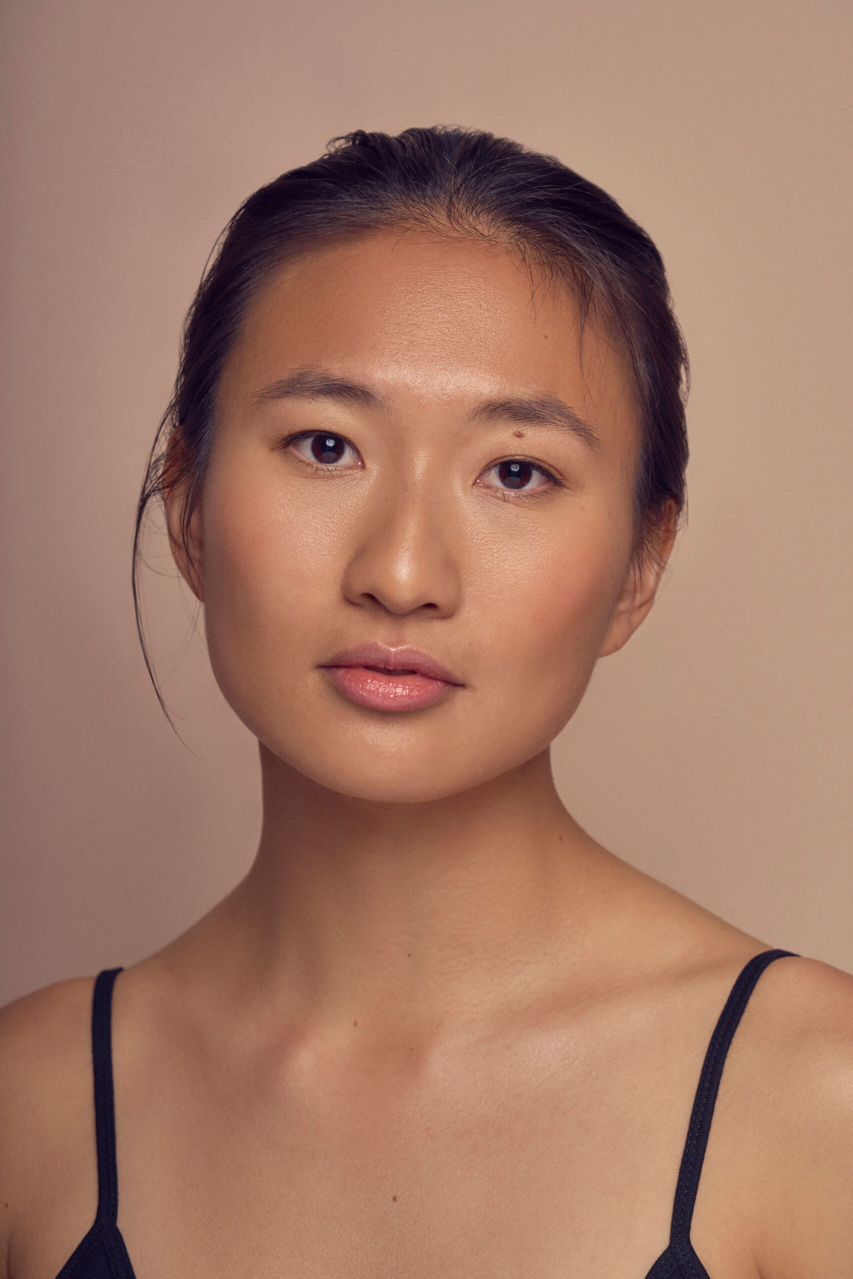 A person with long hair pulled back looks at the camera with a neutral expression. They are wearing a thin-strapped top. The background is a soft beige color.