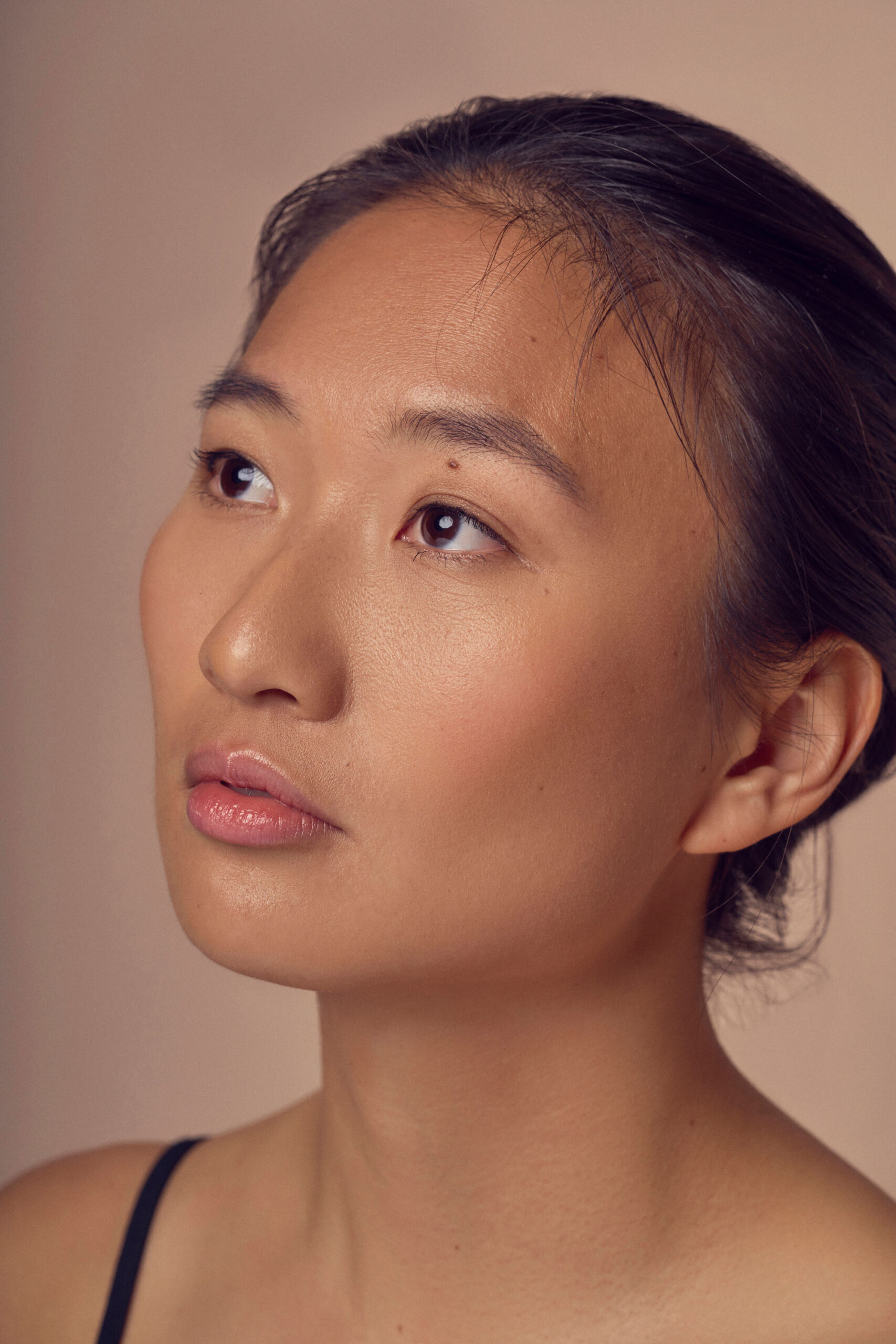 A person with long dark hair pulled back gazes upward. They have smooth, tan skin and are wearing a natural makeup look. The background is a soft beige color.