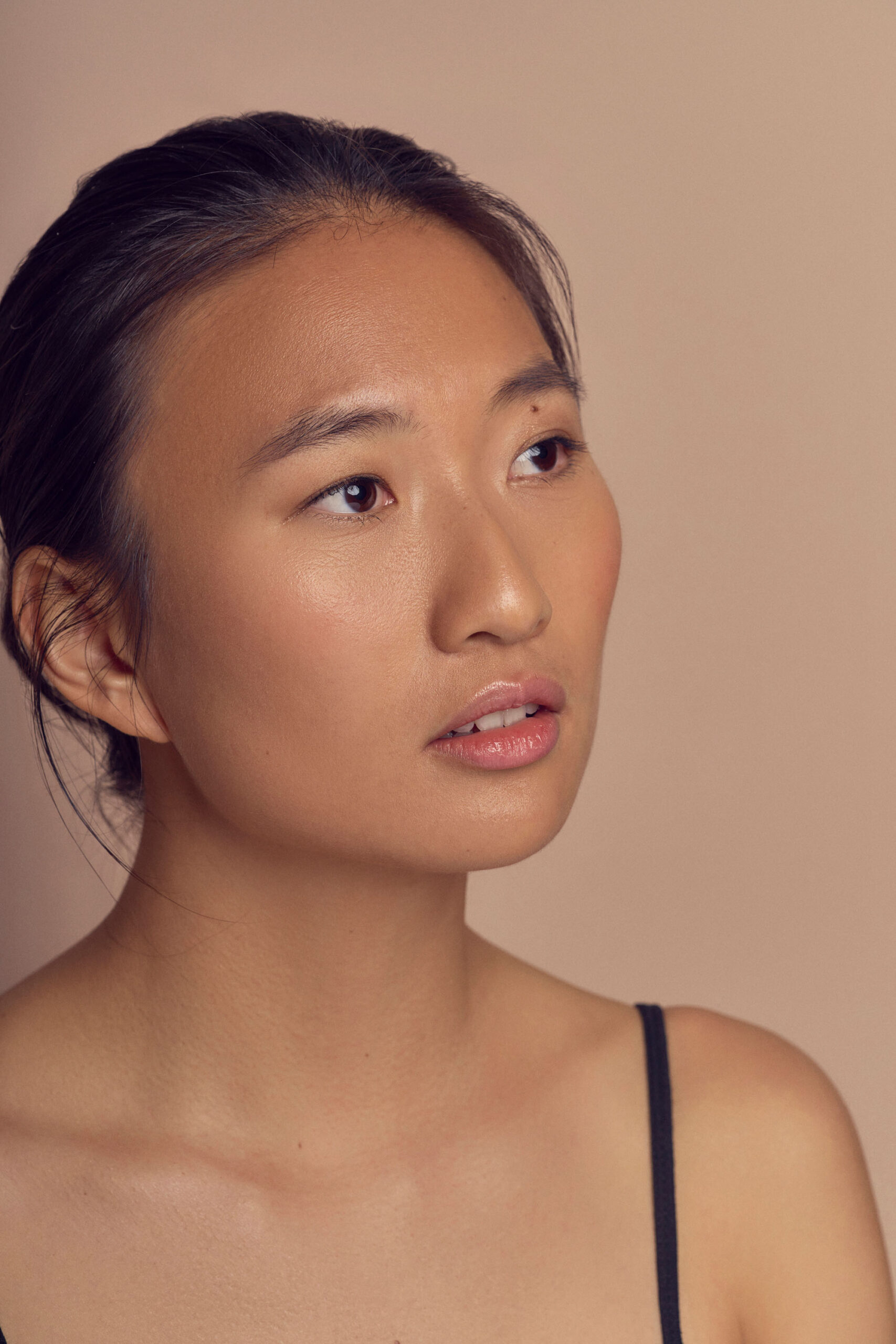 A person with a neutral expression looks upward against a neutral, tan background. They have long dark hair tied back and are wearing a thin-strapped black top. The lighting softly highlights their facial features.