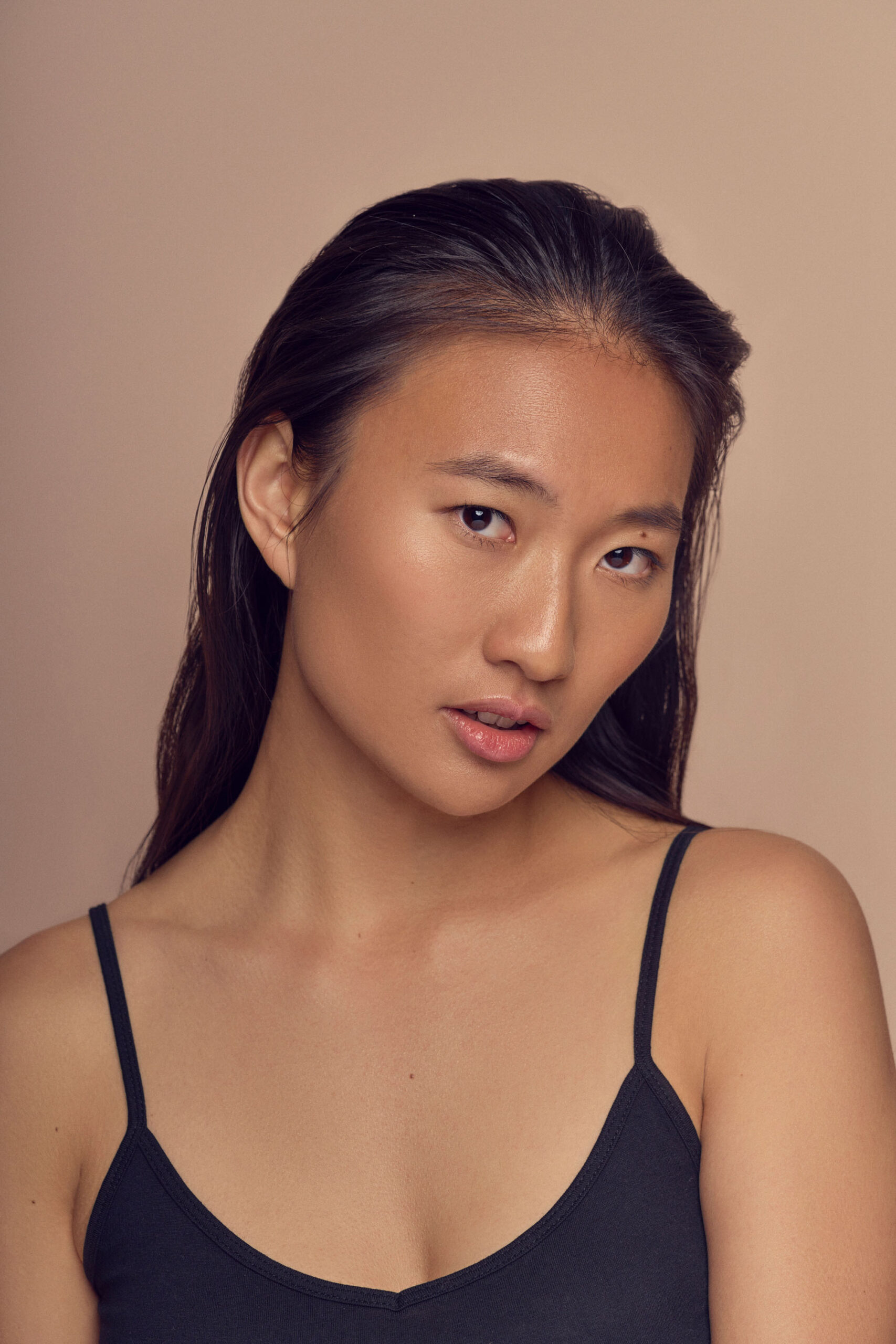 A person with long dark hair wearing a black tank top poses against a beige background. They have a neutral expression and are slightly tilting their head to the side.