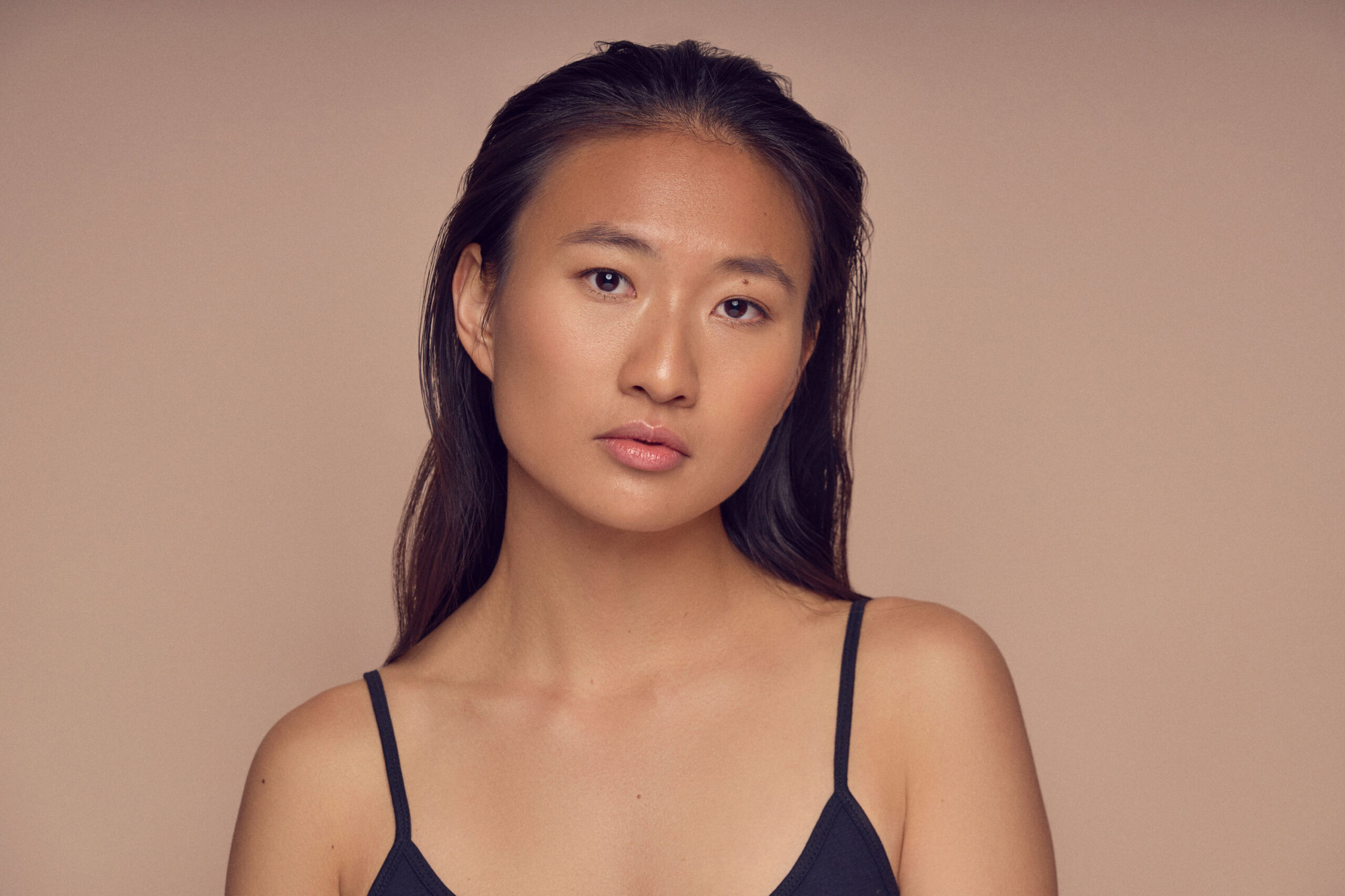 Portrait of a person with long dark hair, wearing a black strap top, against a plain beige background, looking directly at the camera with a neutral expression.