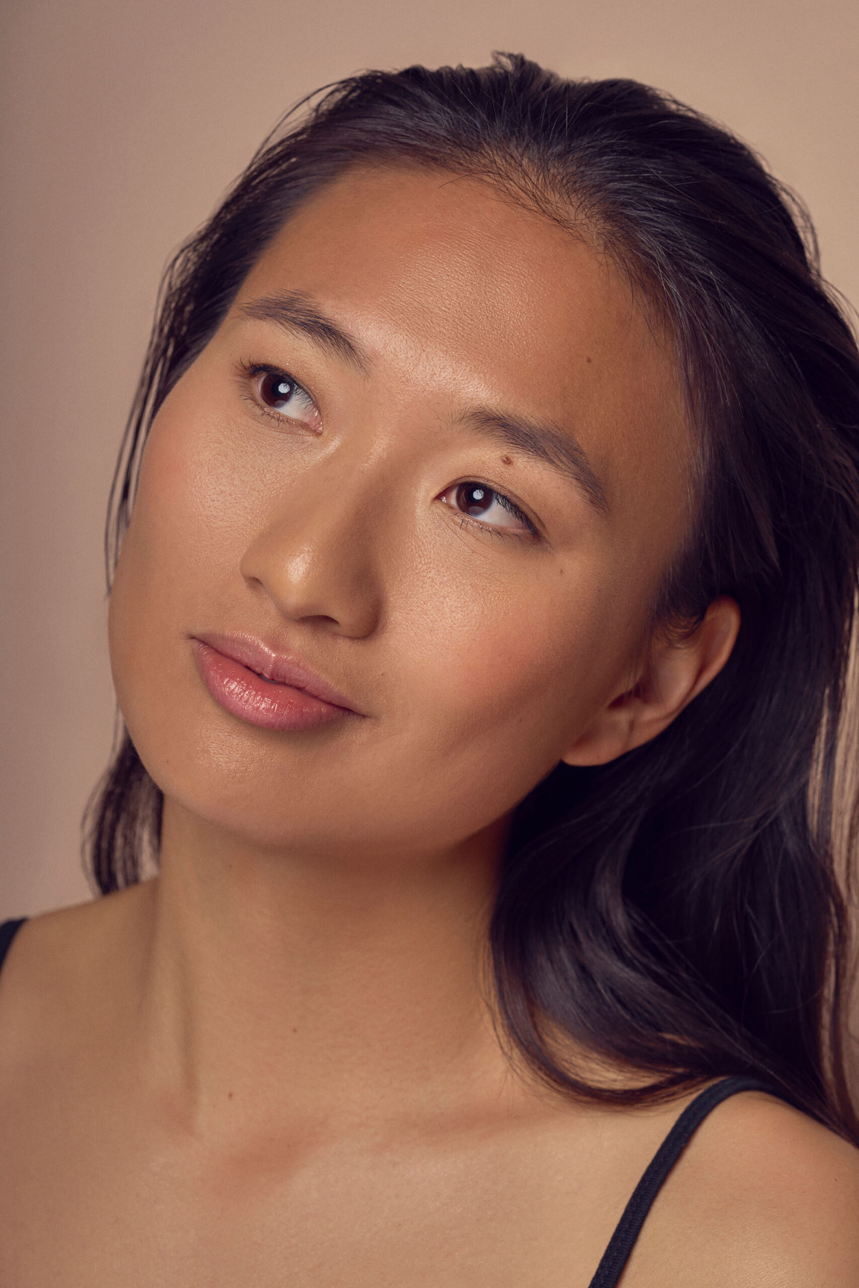 A person with long, dark hair looks upward with a thoughtful expression. The setting has soft, neutral lighting, highlighting their smooth skin and relaxed demeanor. They are wearing a black, sleeveless top.