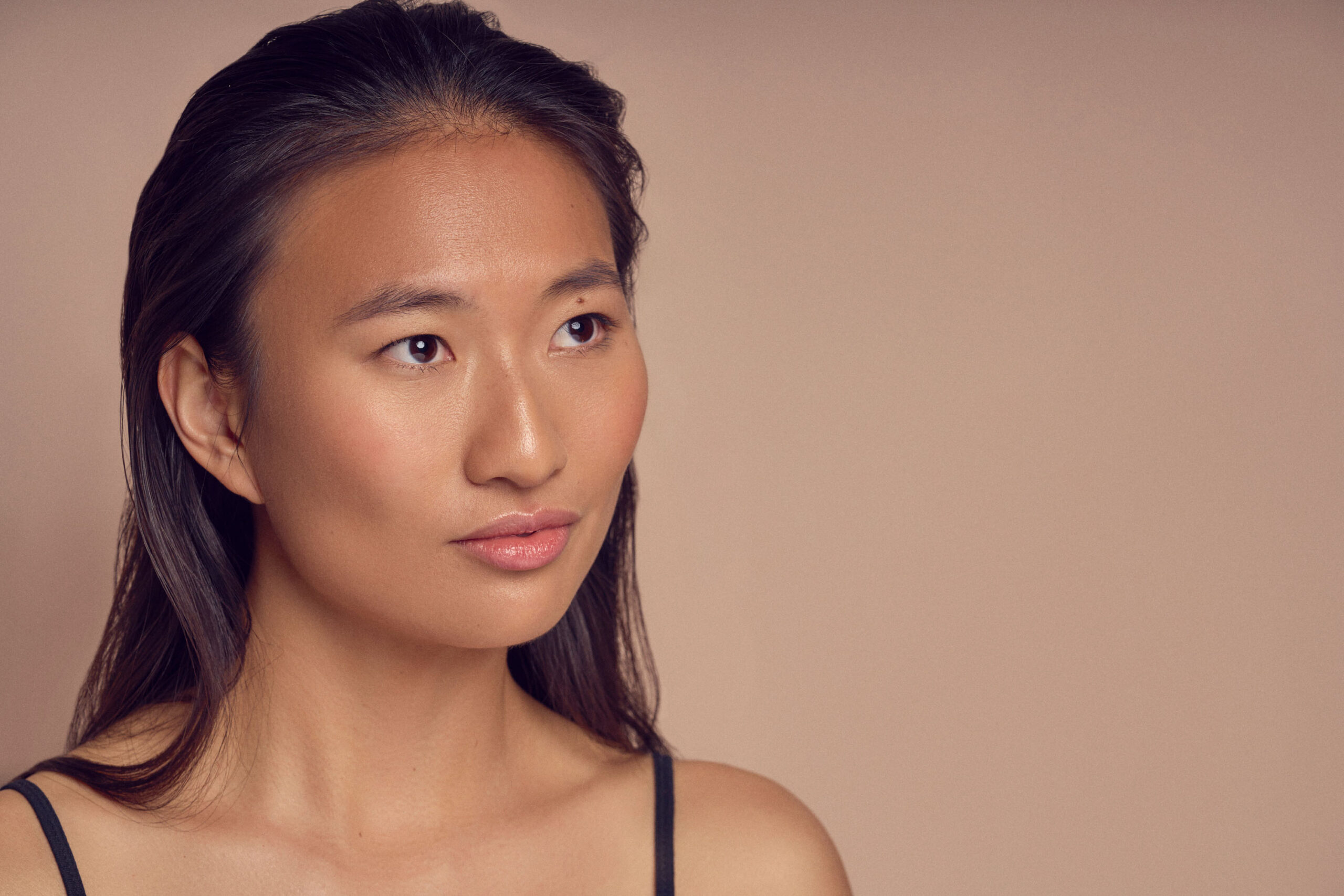 A person with long dark hair gazes to the right, wearing a black strap top. The background is a soft beige color, creating a serene and neutral atmosphere.
