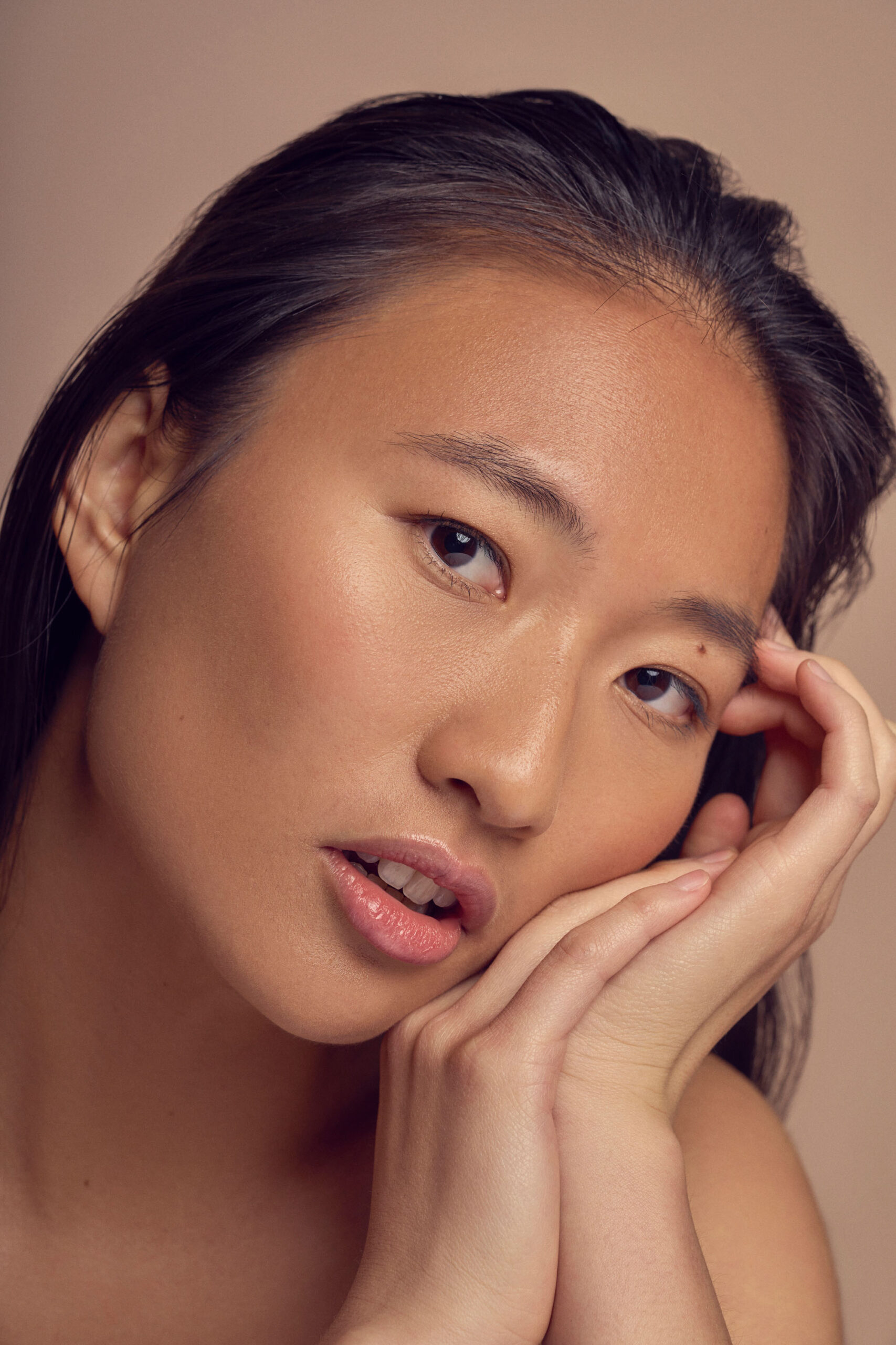 A person with long dark hair poses with their face resting on their hands. They have a neutral expression and are set against a soft, neutral background.