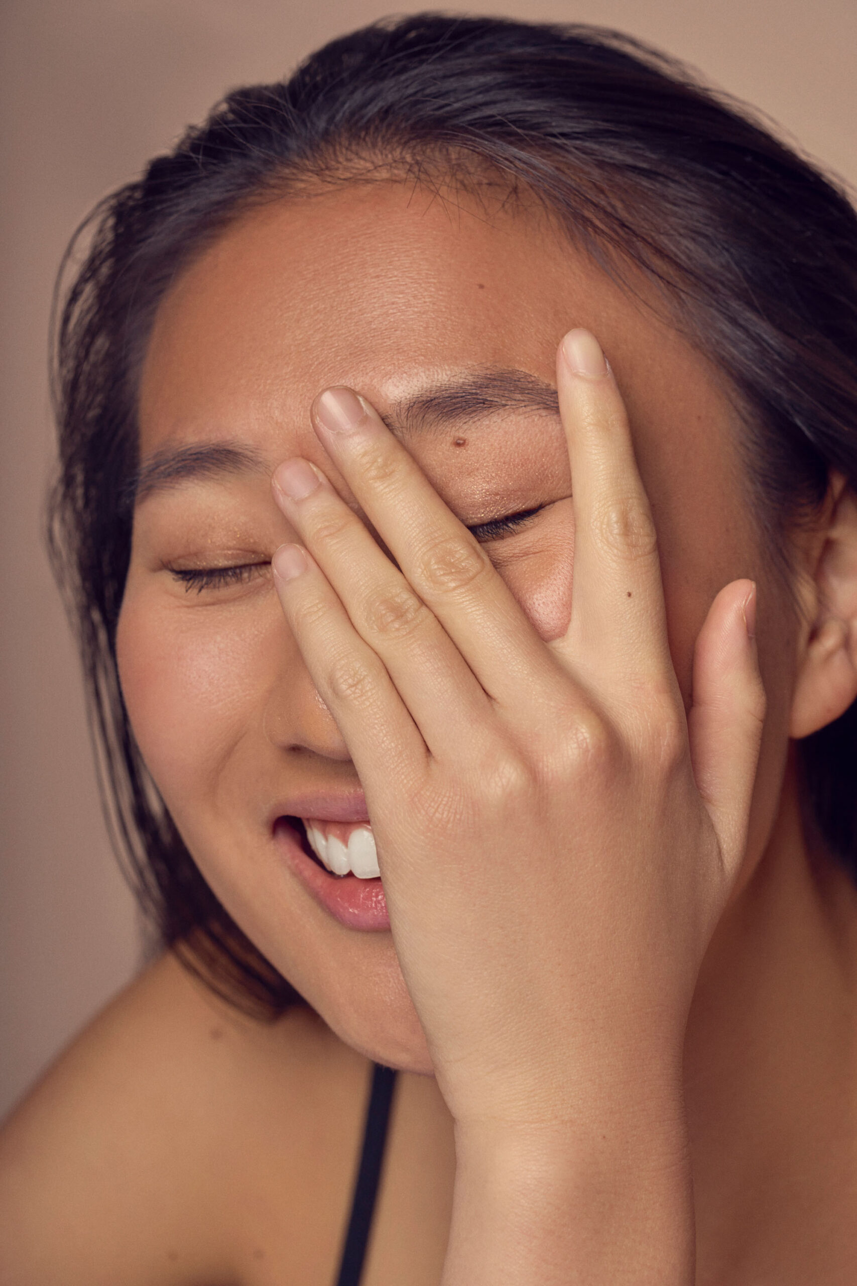 A person with long dark hair smiles with eyes closed, covering part of their face with one hand. They appear joyful and relaxed. The background is softly blurred.