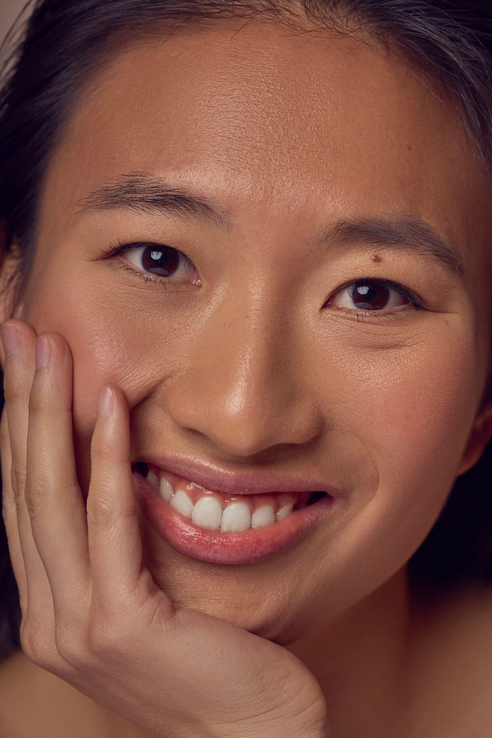 A close-up of a person smiling with their head tilted slightly and resting on their hand. Their eyes are bright and their lips have a natural sheen. The skin has a soft, healthy glow. The background is neutral, drawing focus to their expression.
