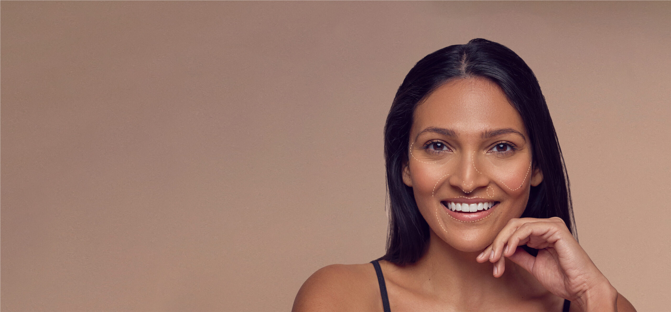 A woman with long dark hair smiles at the camera. Her skin glistens under the light, and she is wearing a sleeveless top. Her hand rests gently under her chin against a smooth beige background.