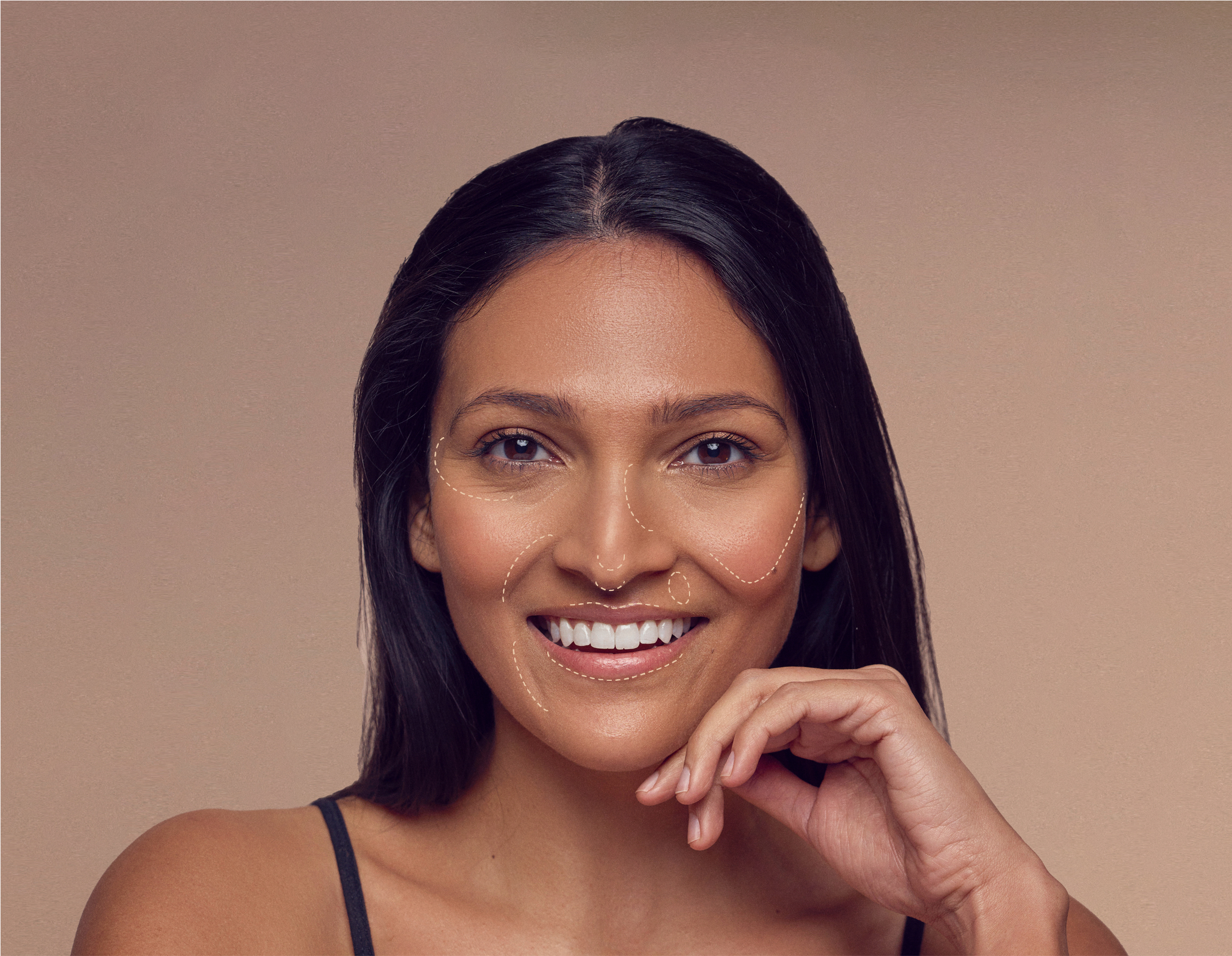 A woman with long dark hair smiles, with contour makeup lines on her face. She rests her chin on her hand against a neutral background.