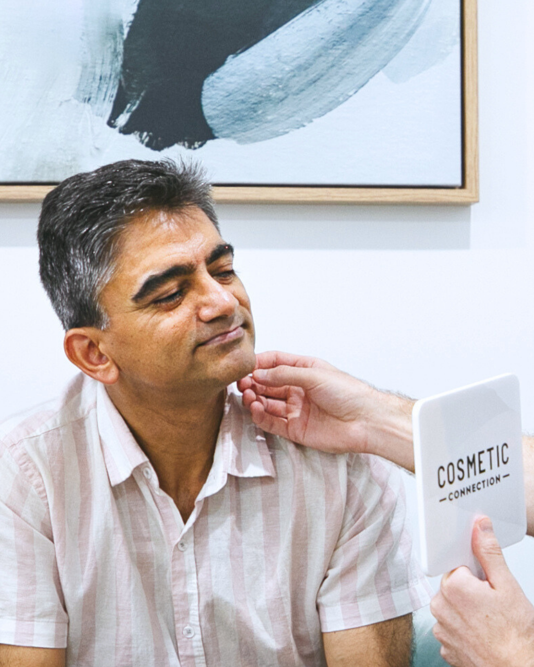 A person in a striped shirt sits while another persons hand gently examines their jaw. They hold a mirror labeled Cosmetic Connection. An abstract painting hangs in the background.